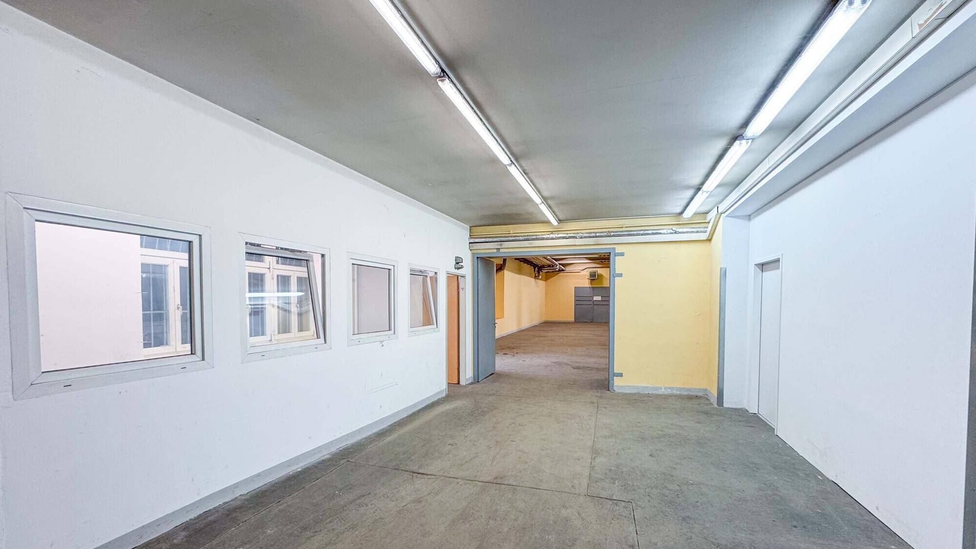 Langer, heller Flur mit Fenstern und Blick in einen weiteren Raum mit gelben Wänden und Betonboden.
