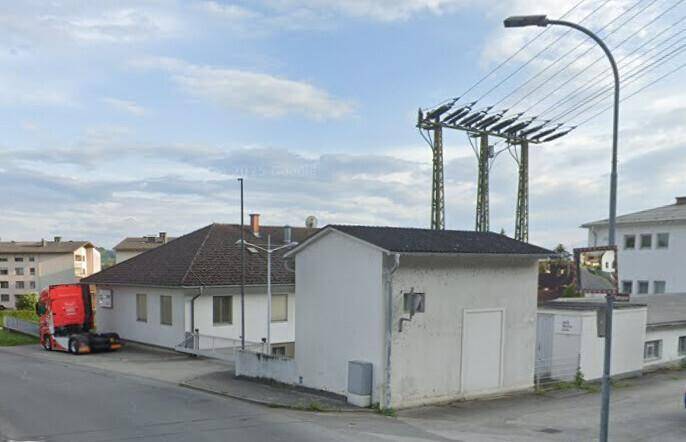 Ansicht eines weißen Gebäudes mit Satteldach, davor ein Lastwagen und Stromleitungen im Hintergrund.
