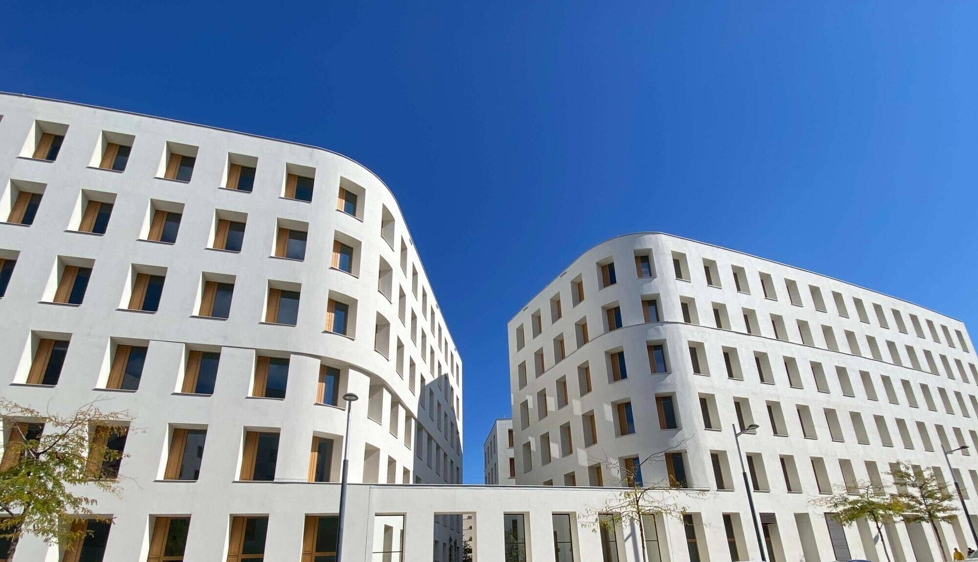 Zwei moderne, weiße Gebäude mit vielen kleinen Fenstern unter blauem Himmel.