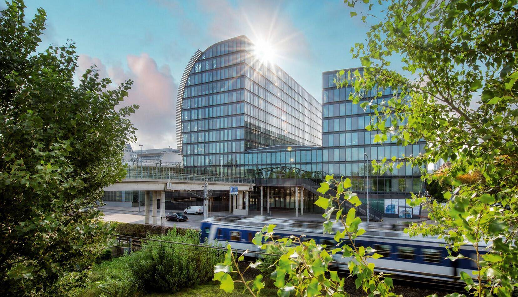 Modernes Bürogebäude mit geschwungener Glasfassade, umgeben von Bäumen und einer Bahntrasse unter strahlendem Himmel.
