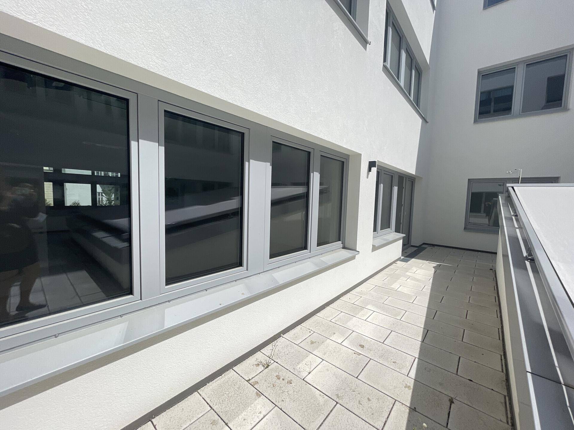 Eine gepflasterte Terrasse entlang der Gebäudefassade mit großen Fenstern und viel Sonnenlicht.