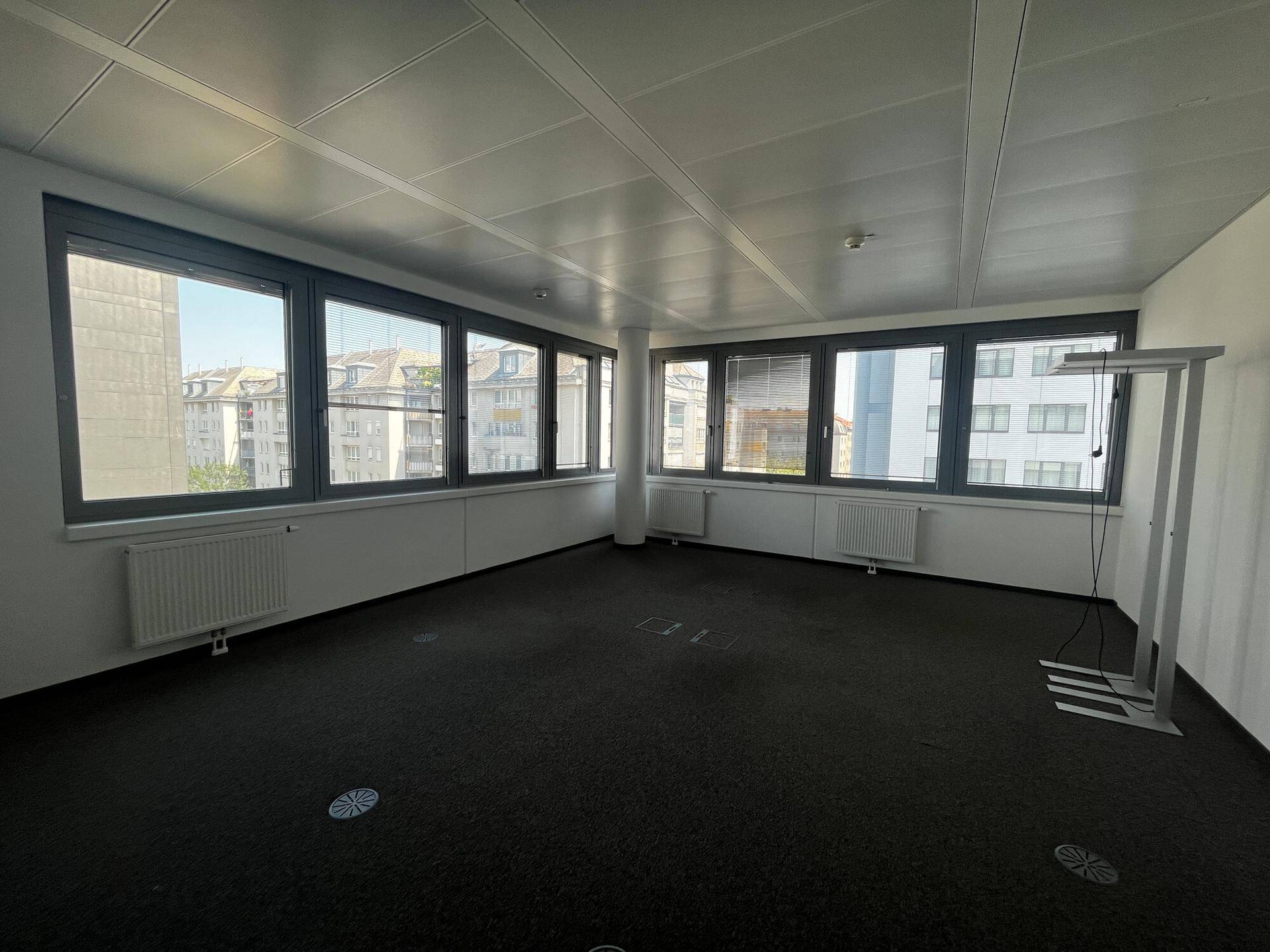 Helles Büro mit Teppichboden, breiter Fensterfront und Blick auf die Stadt, ideal für Arbeitsplätze.