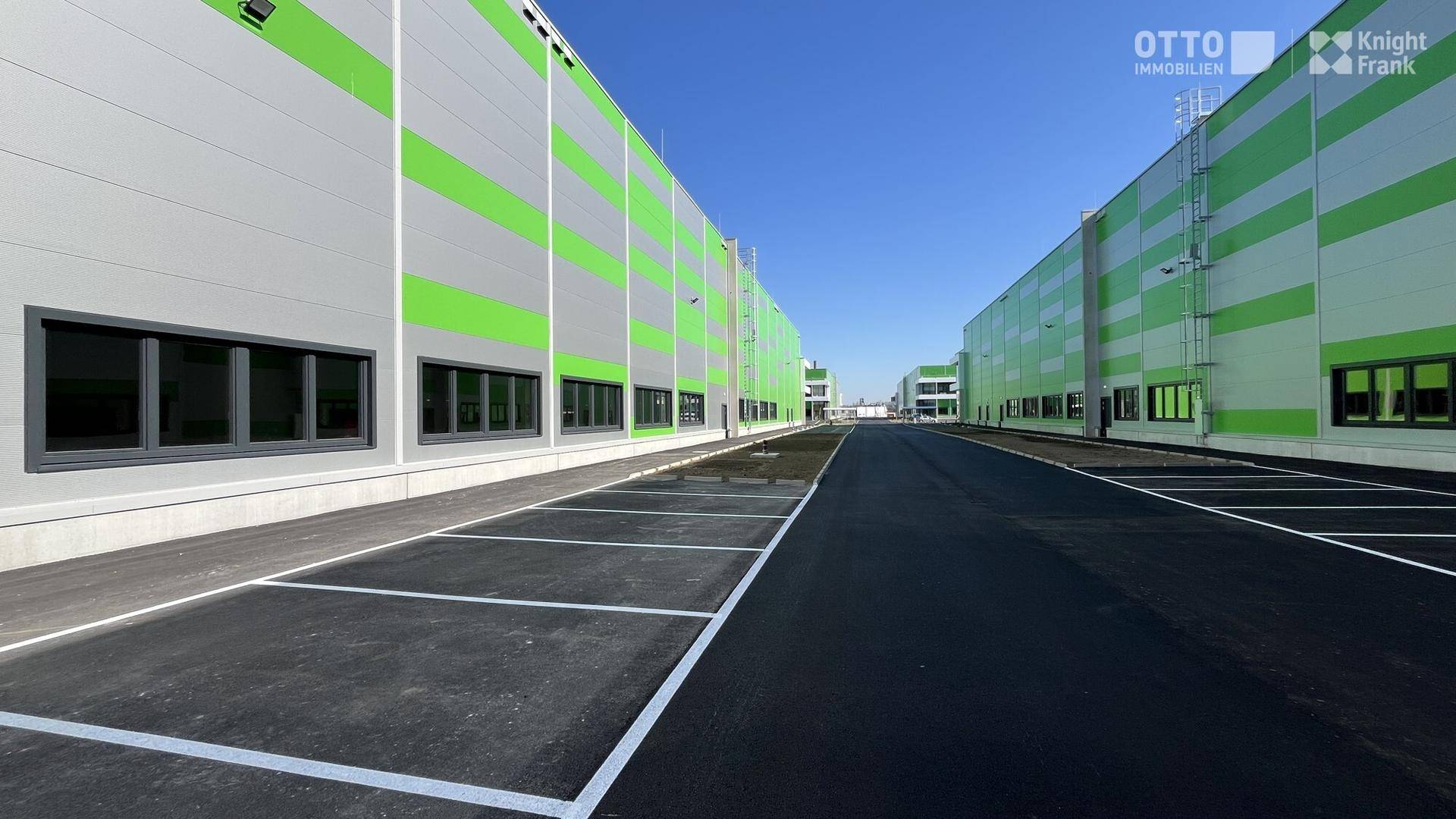 Seitenansicht eines modernen Industriegebäudes mit markanten grün-grauen Streifen und angrenzenden Parkbuchten.