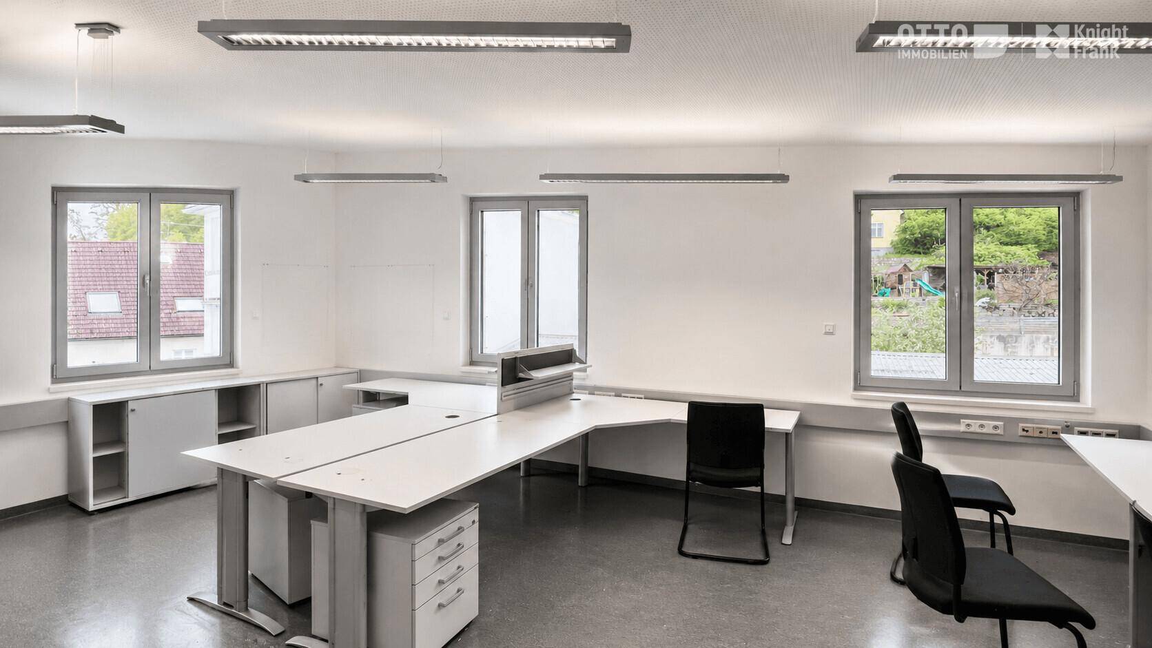Heller Büroraum mit mehreren Arbeitsplätzen, Schreibtischen, Bürostühlen und großen Fenstern.