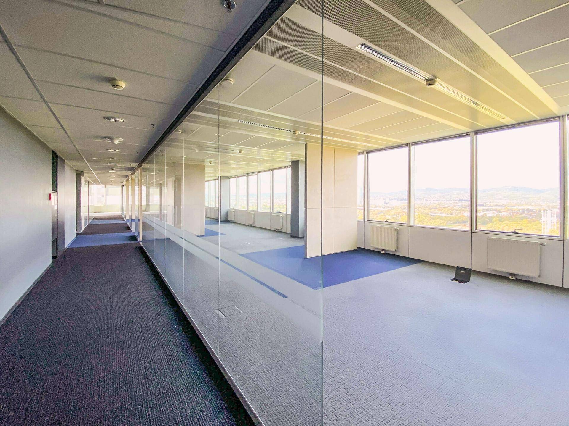 Langer Bürogang mit Glaswänden, der zu einem hellen Großraumbüro mit Stadtblick führt.