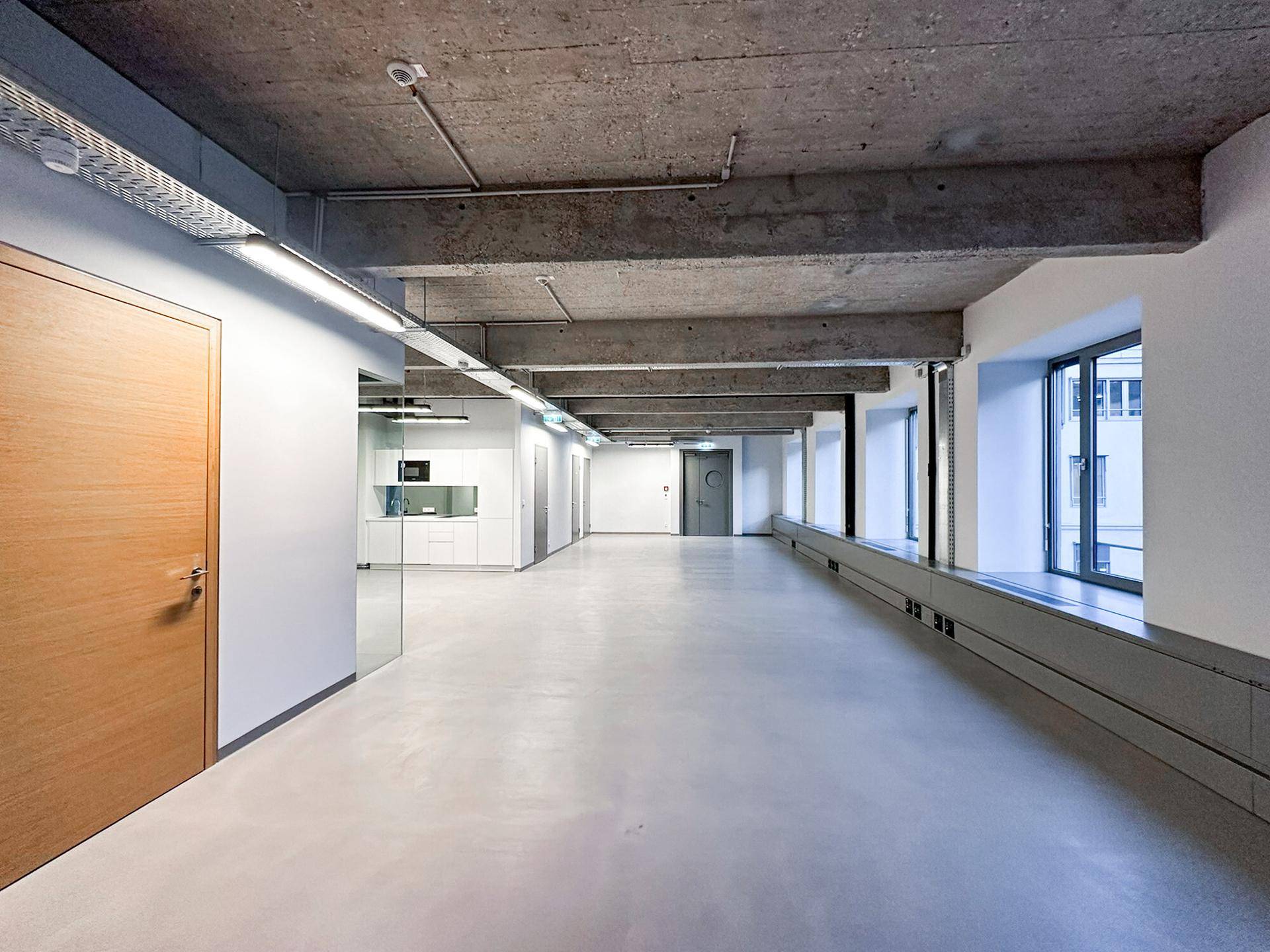 Langer, offener Bürobereich mit Holztüren, hohen Decken und einer Küchenzeile im Hintergrund, ideal für Großraumbüros.