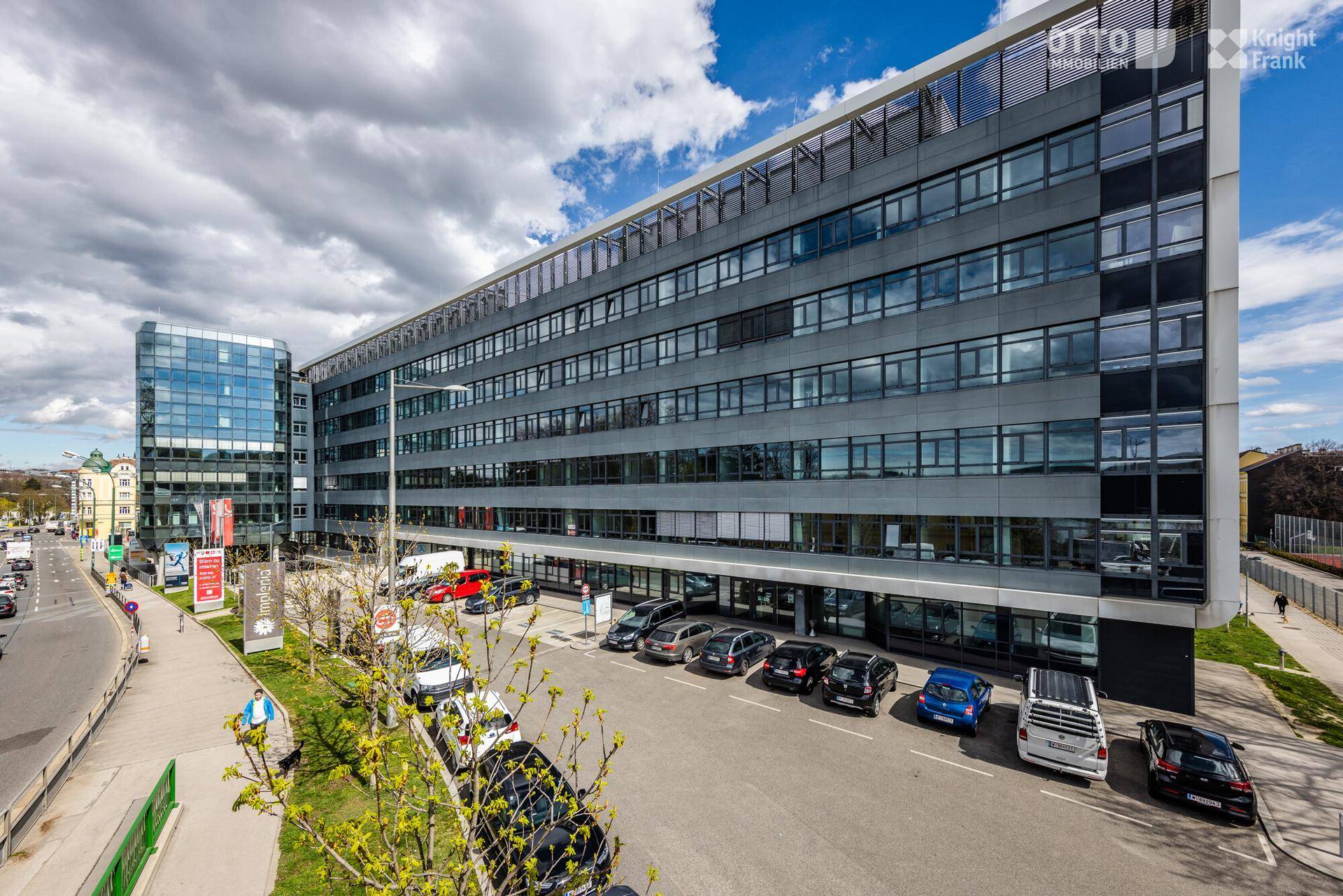 Modernes Bürogebäude mit Glasfassade und zahlreichen Fenstern, umgeben von einem Parkplatz unter bewölktem Himmel.