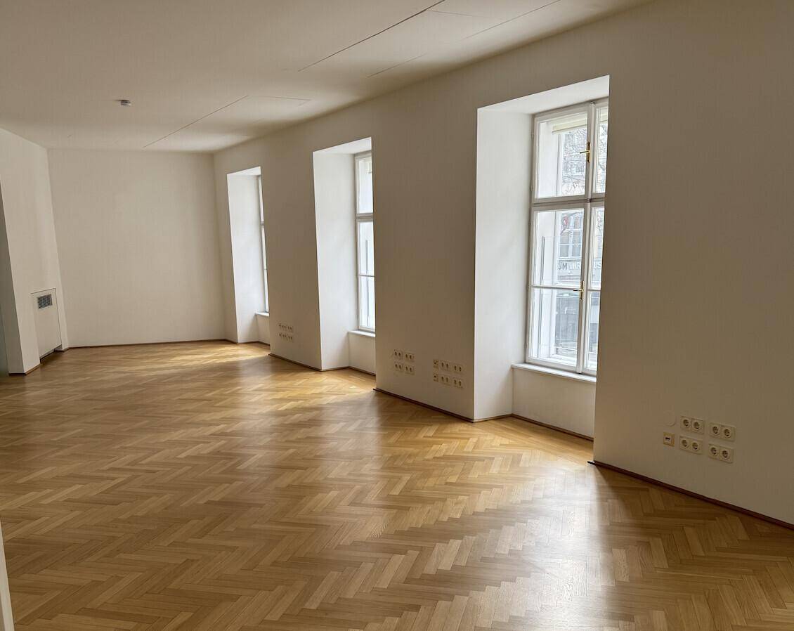 Heller, leerer Raum mit mehreren hohen Fenstern und Fischgrätparkett, bietet viel Tageslicht und Raumgefühl.