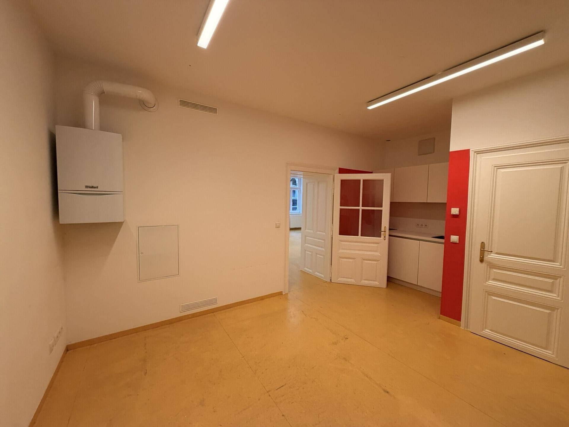 Leerer Raum mit Wandheizkessel und Durchgang zu einer Küchenzeile, ideal für gewerbliche Nutzung.