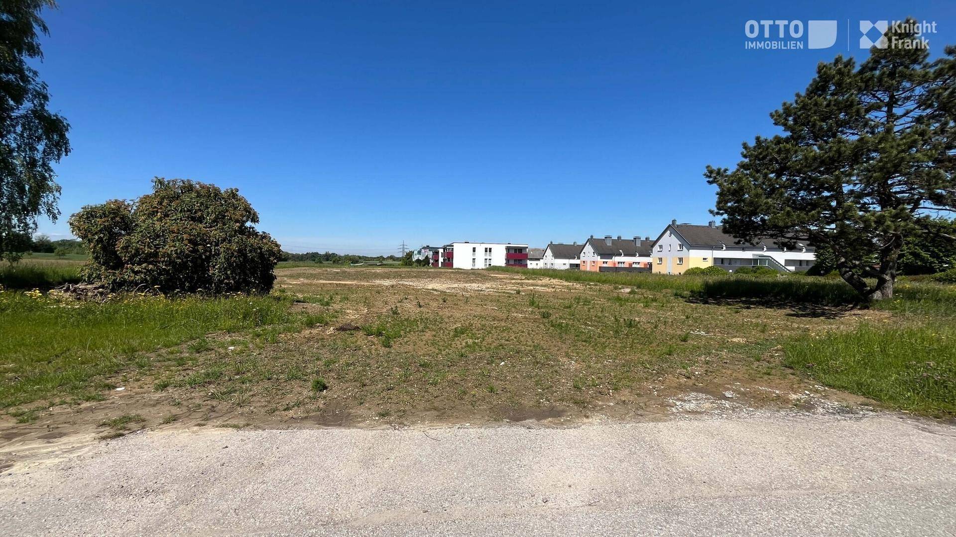 Blick vom Boden auf das weitläufige Baugrundstück mit spärlicher Vegetation und Häusern im Hintergrund unter blauem Himmel.