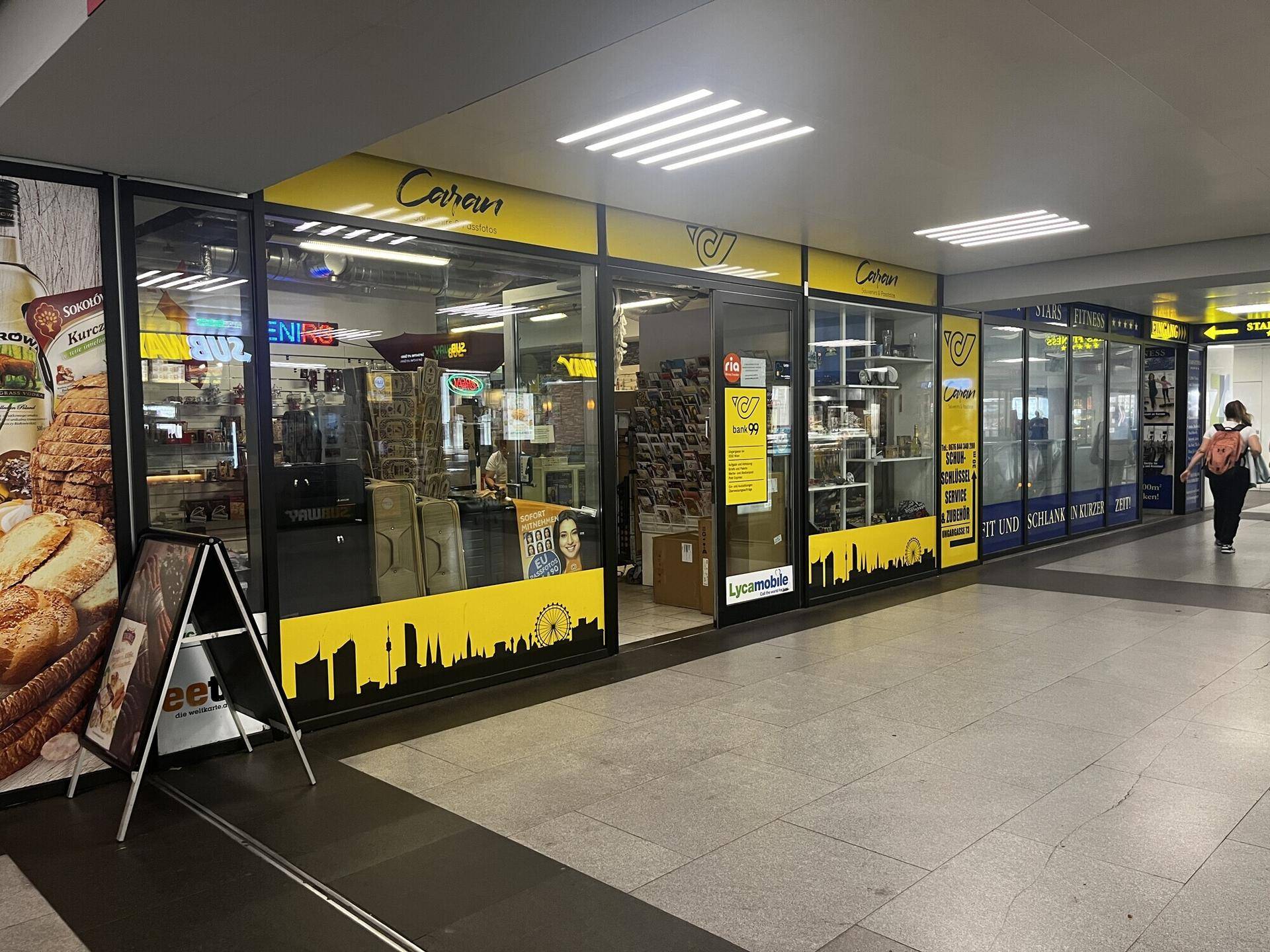Ladenzeile in einem Einkaufszentrum mit verschiedenen Geschäften, großen Schaufenstern und heller Beleuchtung.