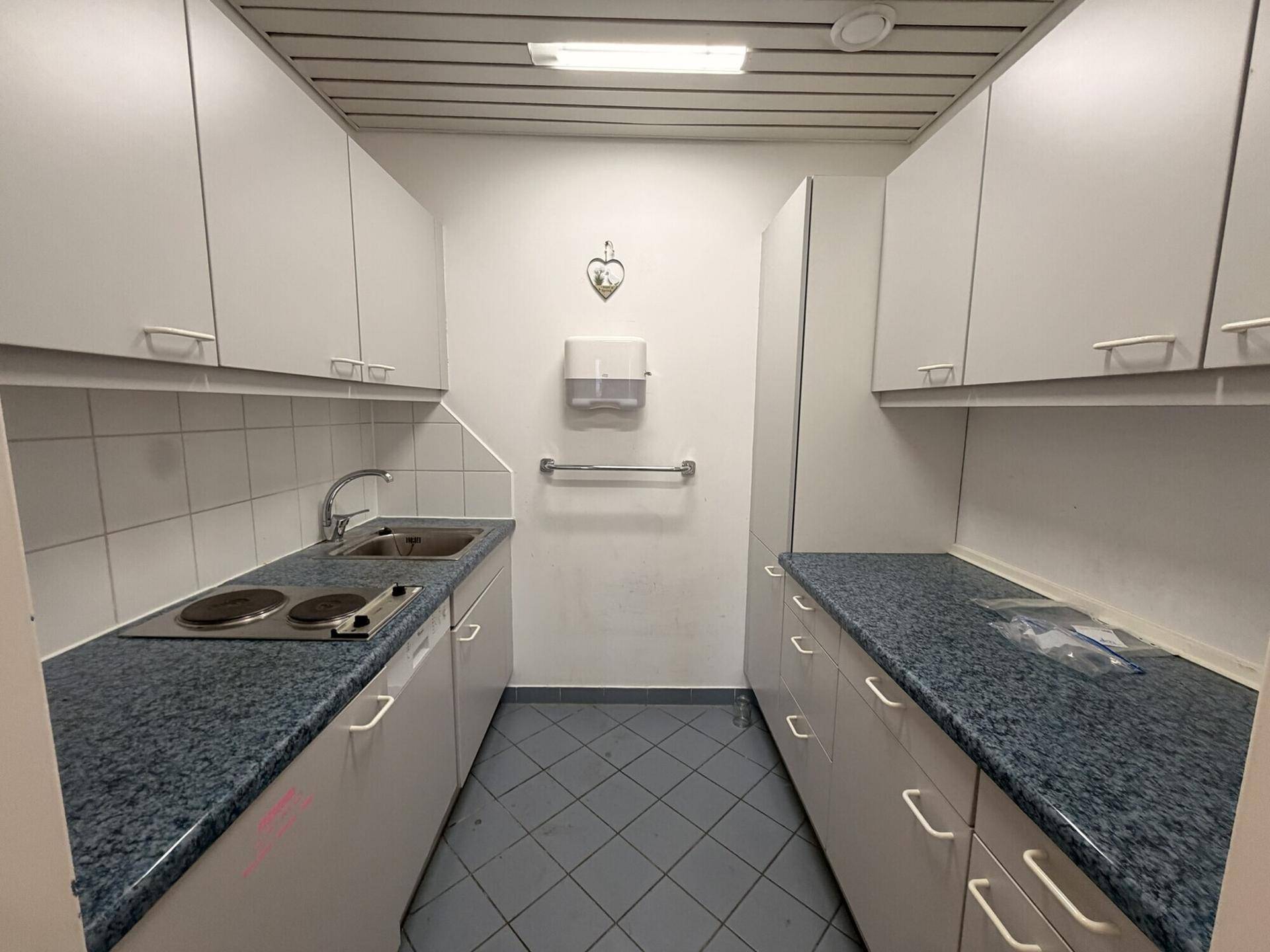 Kleine, funktionale Teeküche mit weißen Schränken, Spüle und Kochplatten, ideal für Pausen.