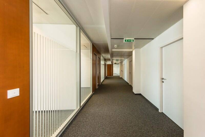 Langer Bürogang mit Teppichboden, Holztüren und Glaswänden, die zu den Büroräumen führen.