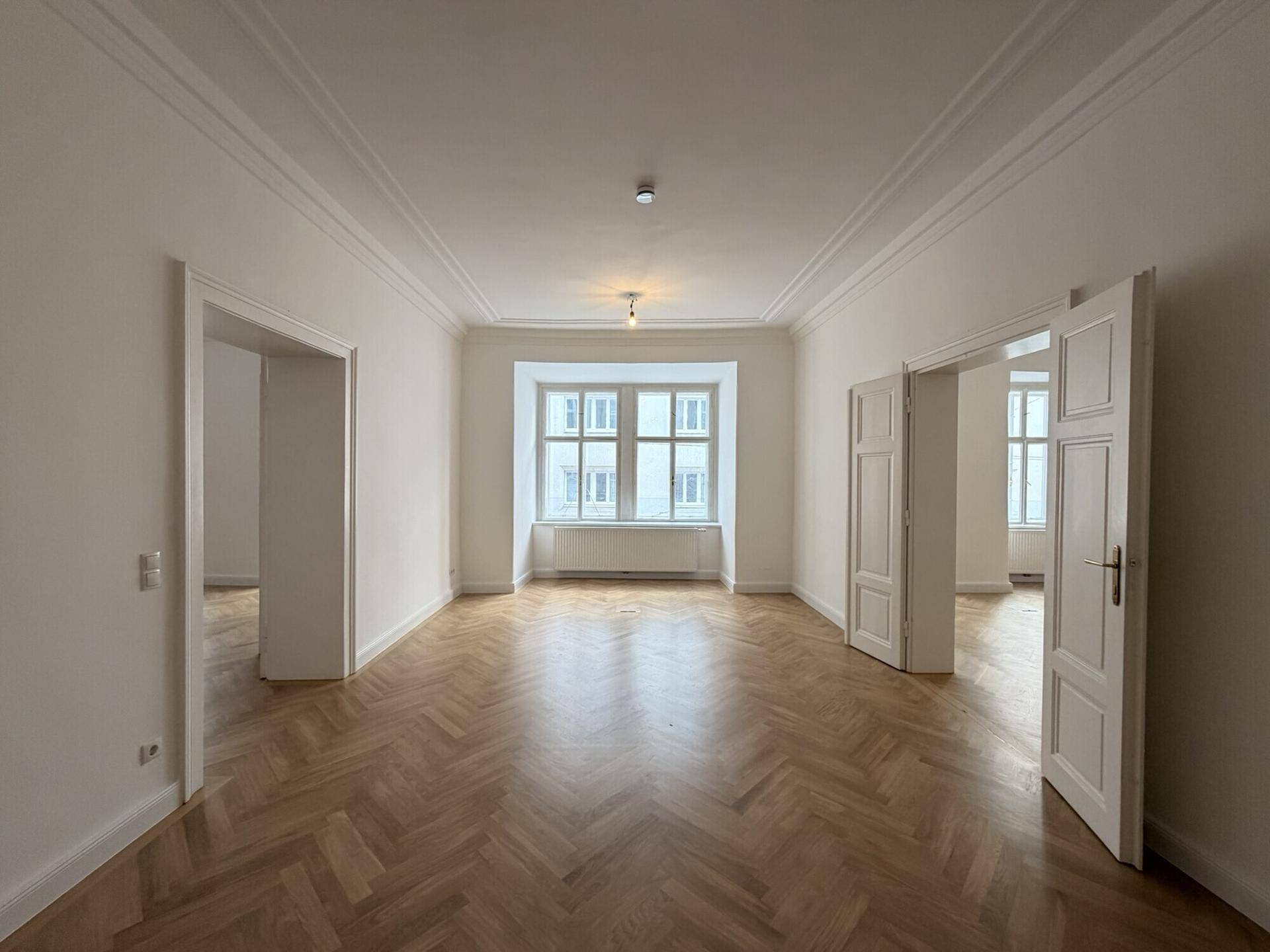 Elegantes Wohnzimmer mit Fischgrätparkett, Stuckdecken und einem großen Fenster, das den Raum erhellt.