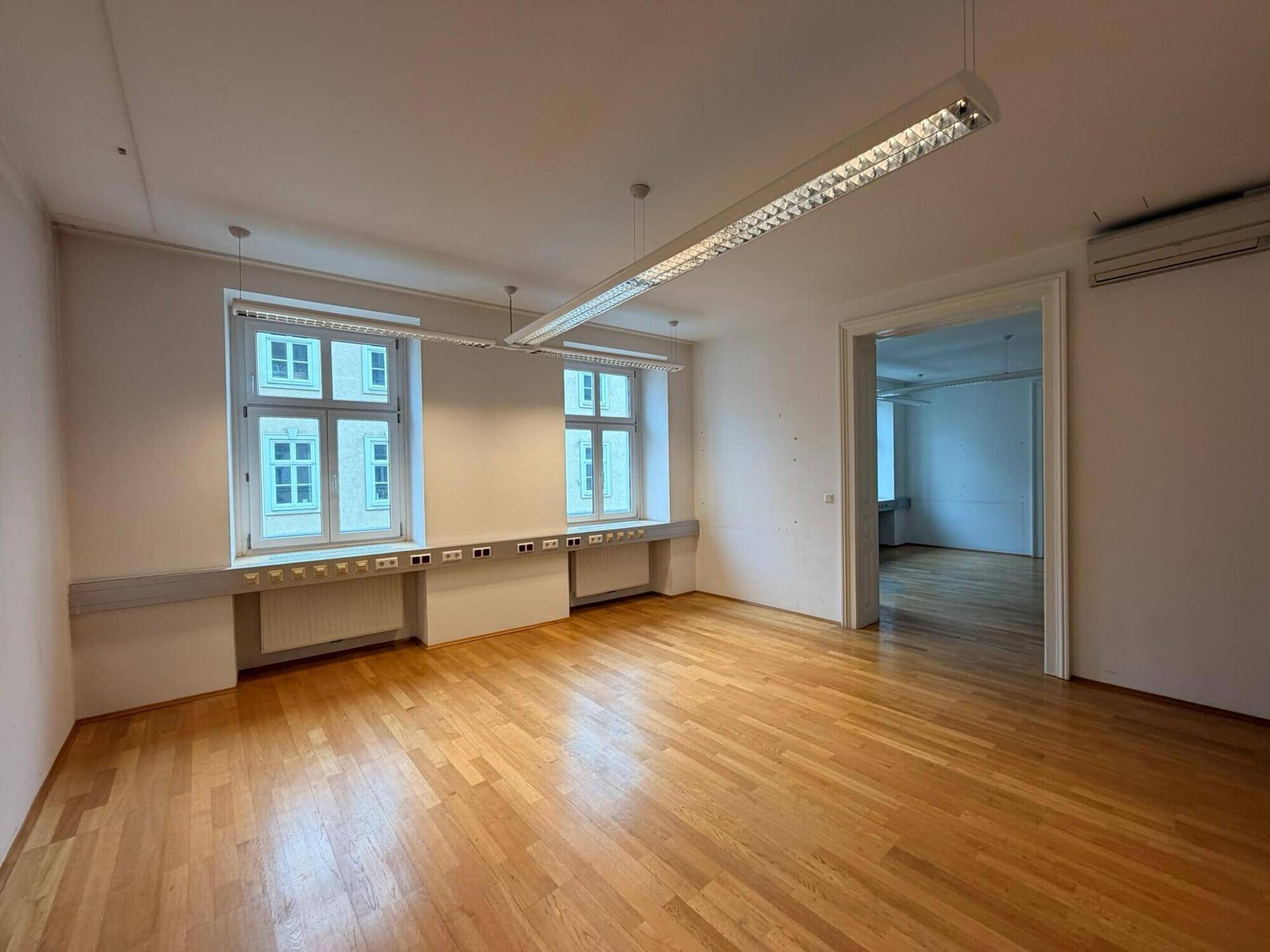 Heller Büroraum mit Holzboden, großen Fenstern und integrierten Arbeitsflächen unter den Fenstern, ideal für Arbeitsplätze.