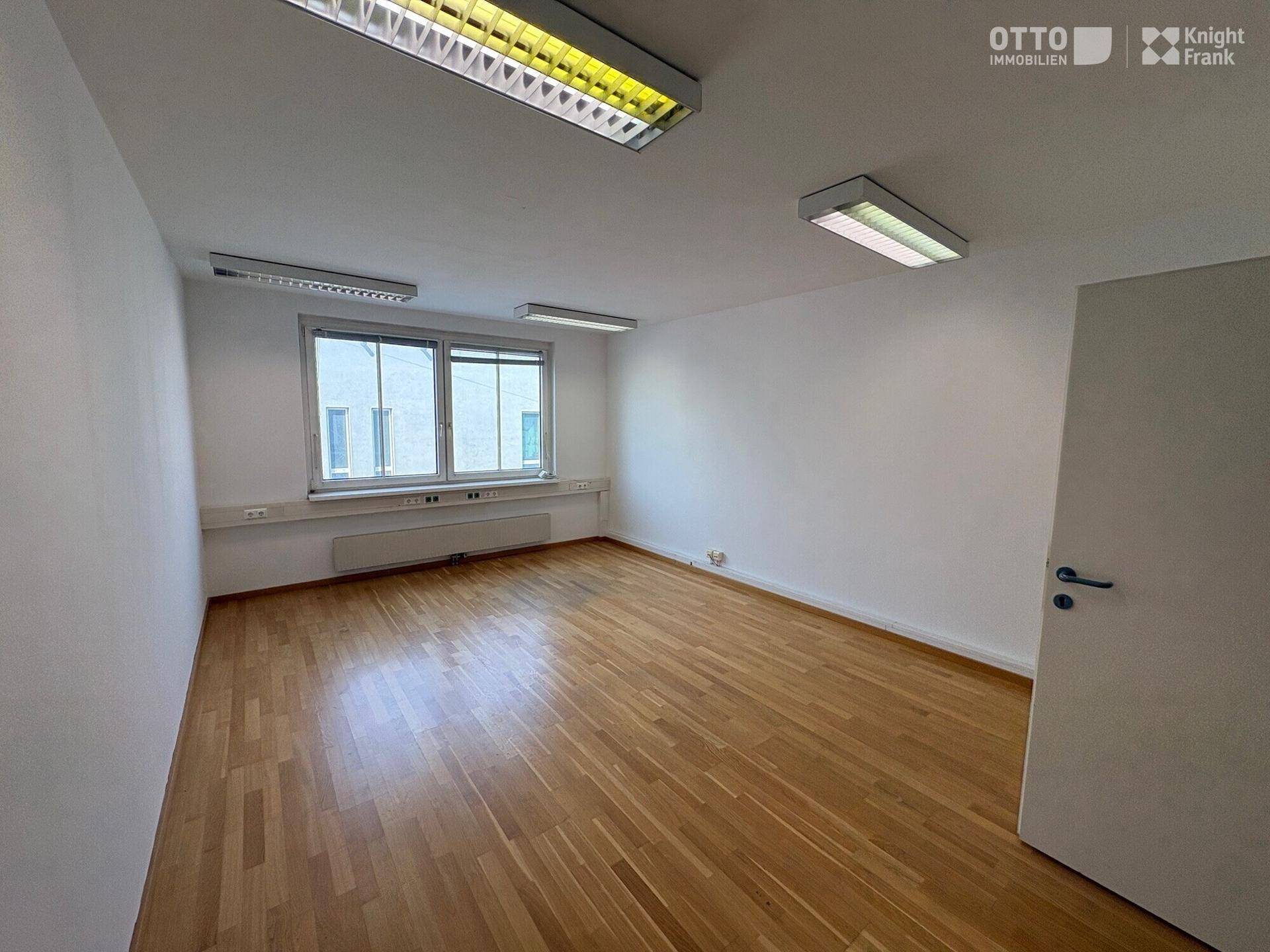 Helles, leeres Büro mit Holzfußboden, großen Fenstern und Deckenbeleuchtung.