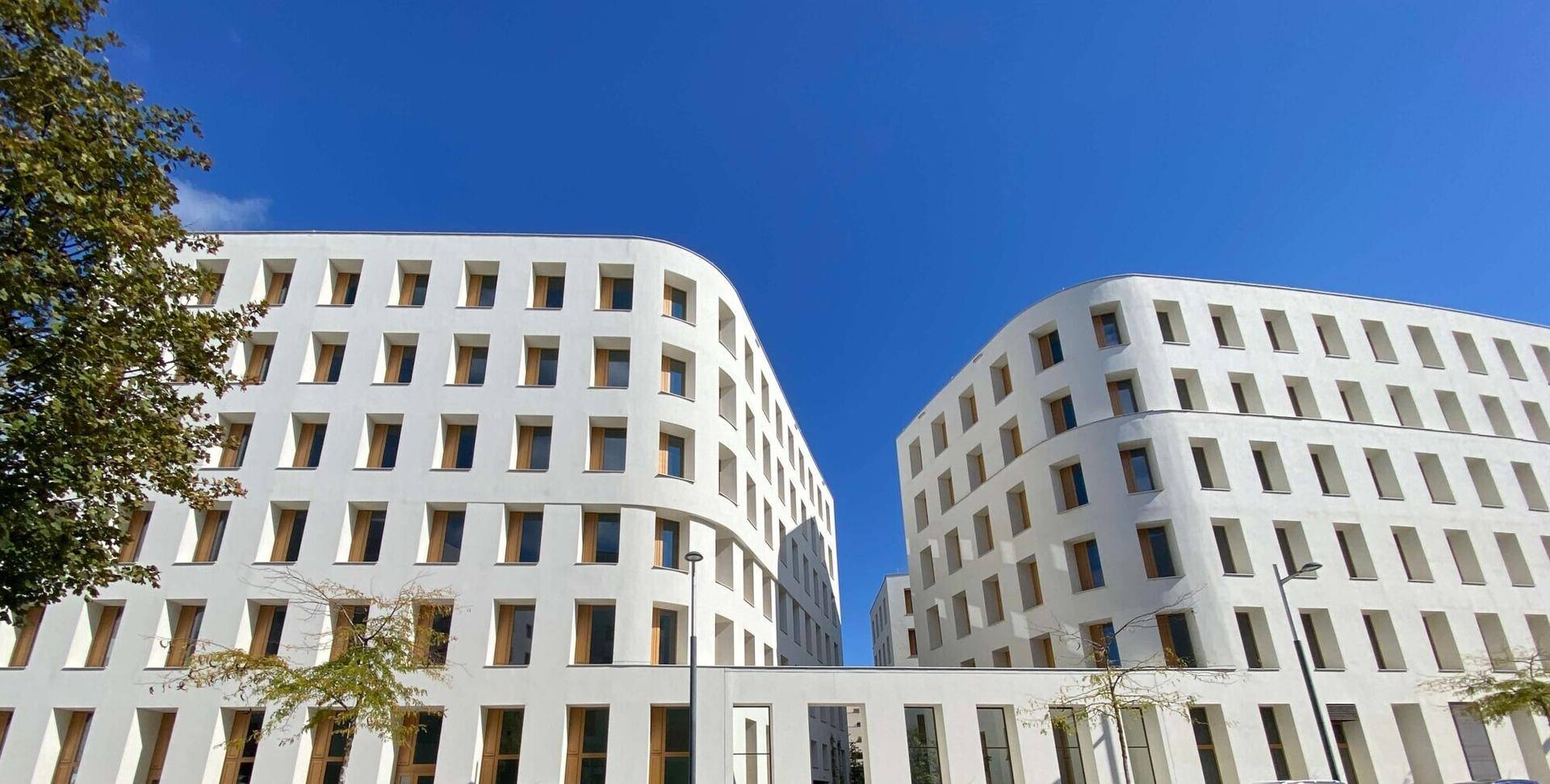 Zwei moderne weiße Gebäude mit geschwungenen Fassaden und zahlreichen Fenstern unter einem strahlend blauen Himmel.