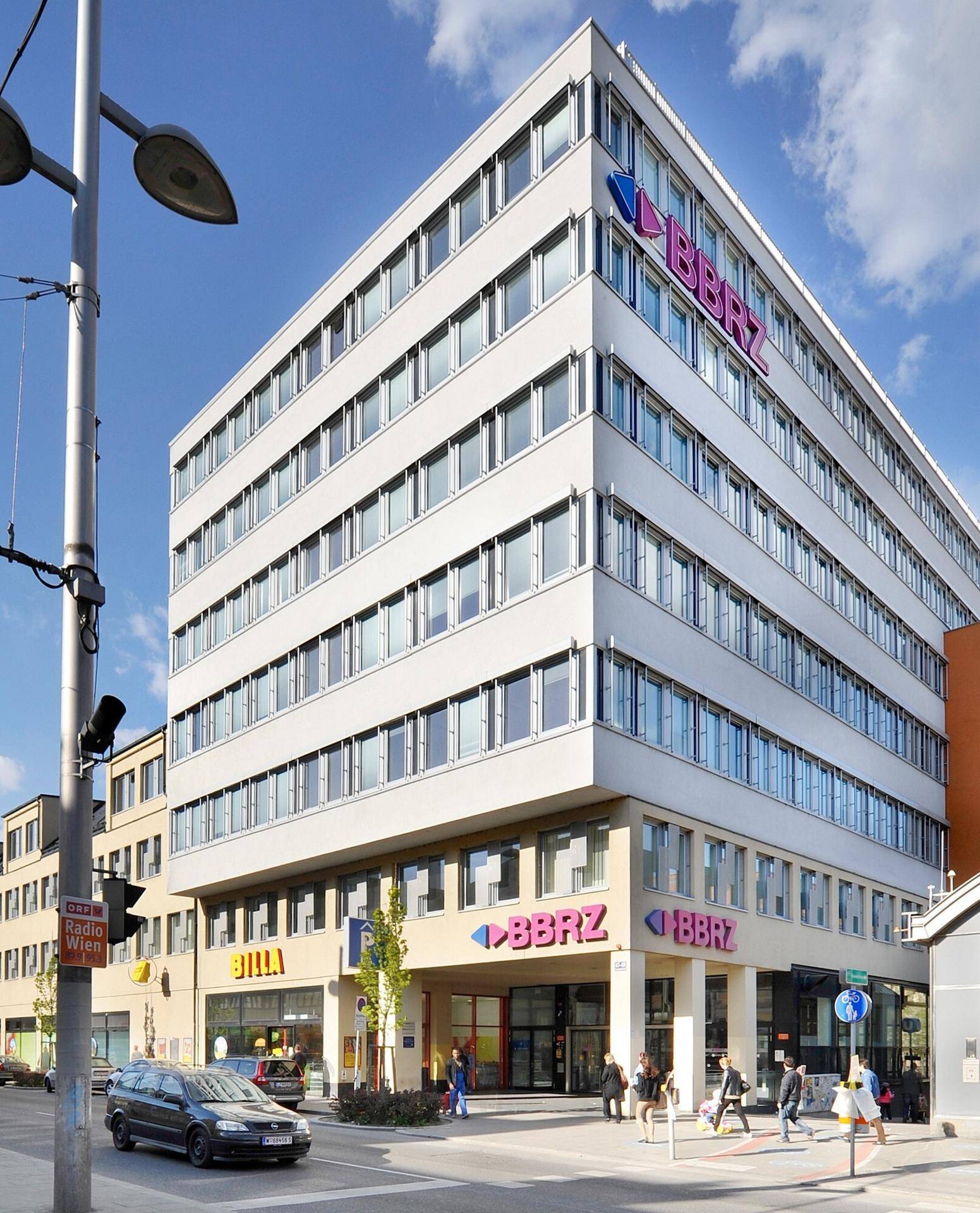 Modernes Bürogebäude an einer belebten Kreuzung mit Geschäften im Erdgeschoss und klarem Himmel.
