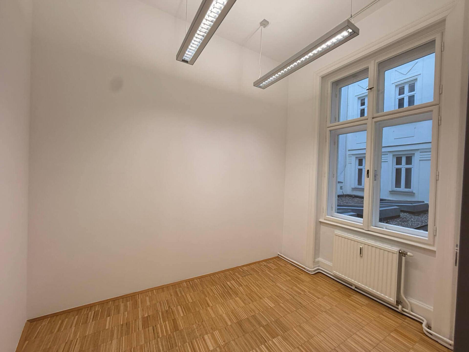 Kompakter Büroraum mit Parkettboden, großem Fenster und moderner Deckenbeleuchtung, ideal für ein Einzelbüro.