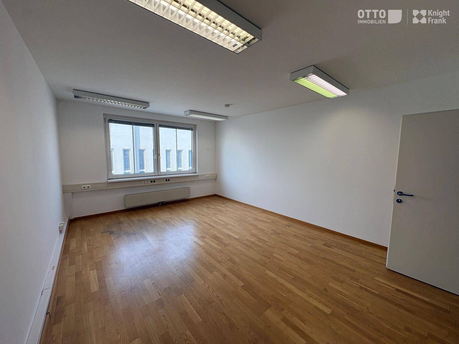 Heller Büroraum mit Holzfußboden, großen Fenstern und effizienter Deckenbeleuchtung.