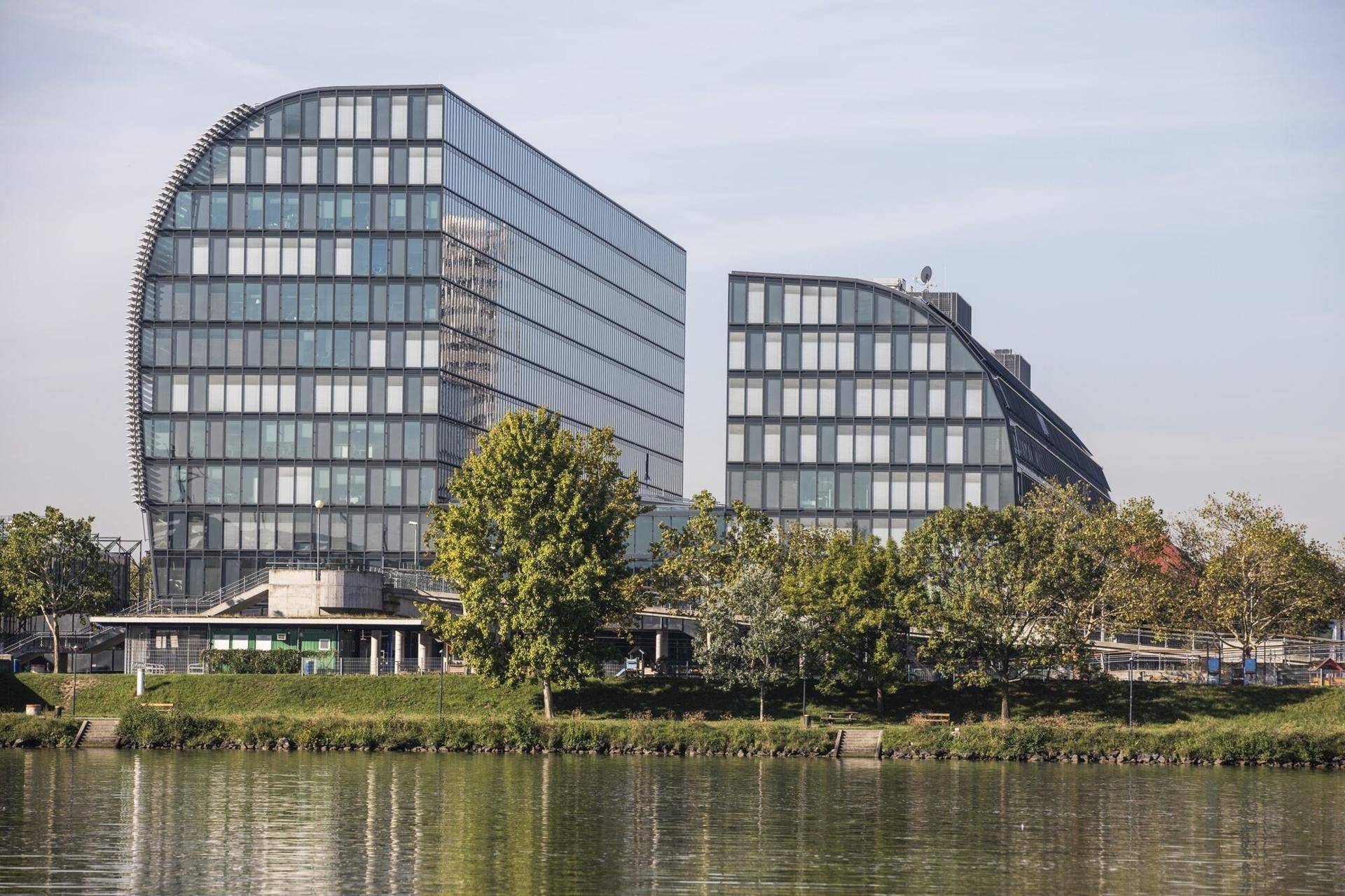 Zwei moderne Bürogebäude mit geschwungenen Glasfassaden am Ufer eines Flusses, umgeben von Bäumen.