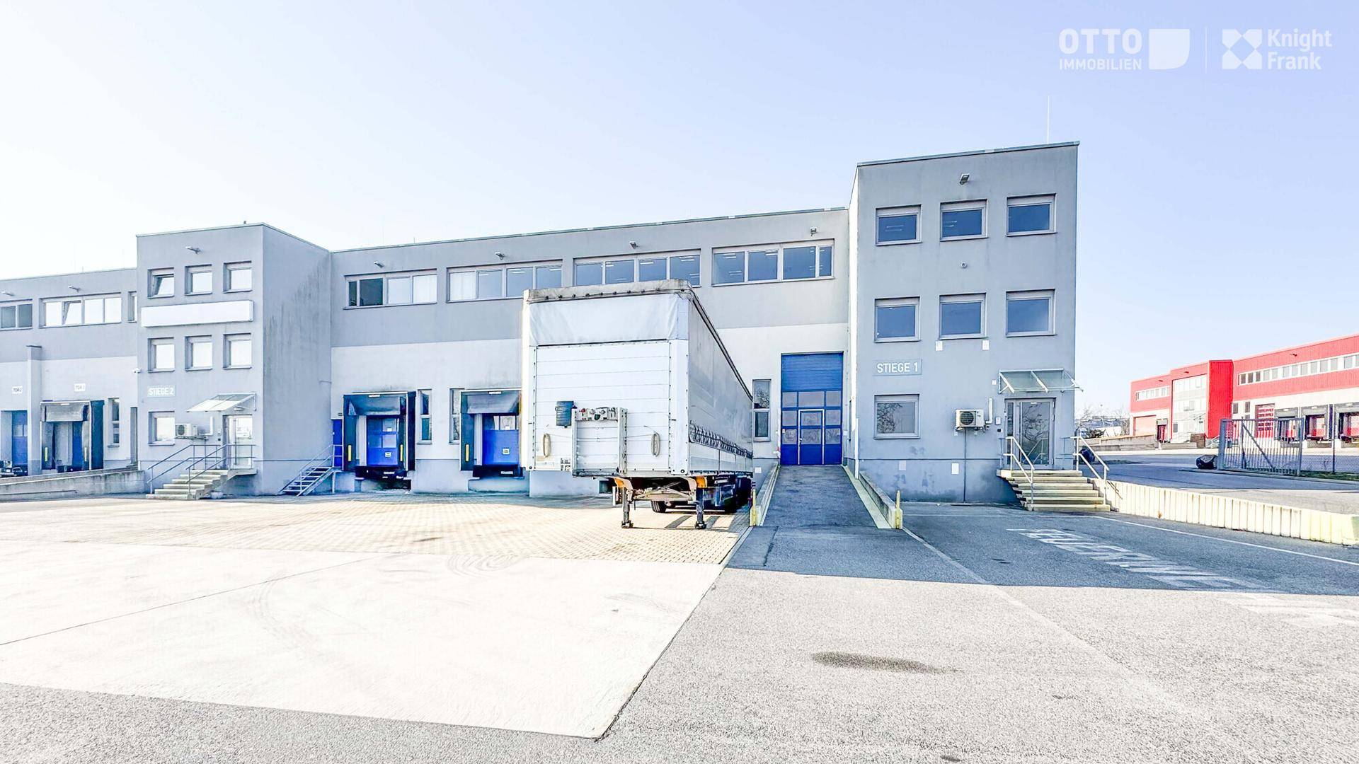 Gewerbegebäude mit Laderampen und einem geparkten Lastwagenanhänger, umgeben von einer großen Freifläche.