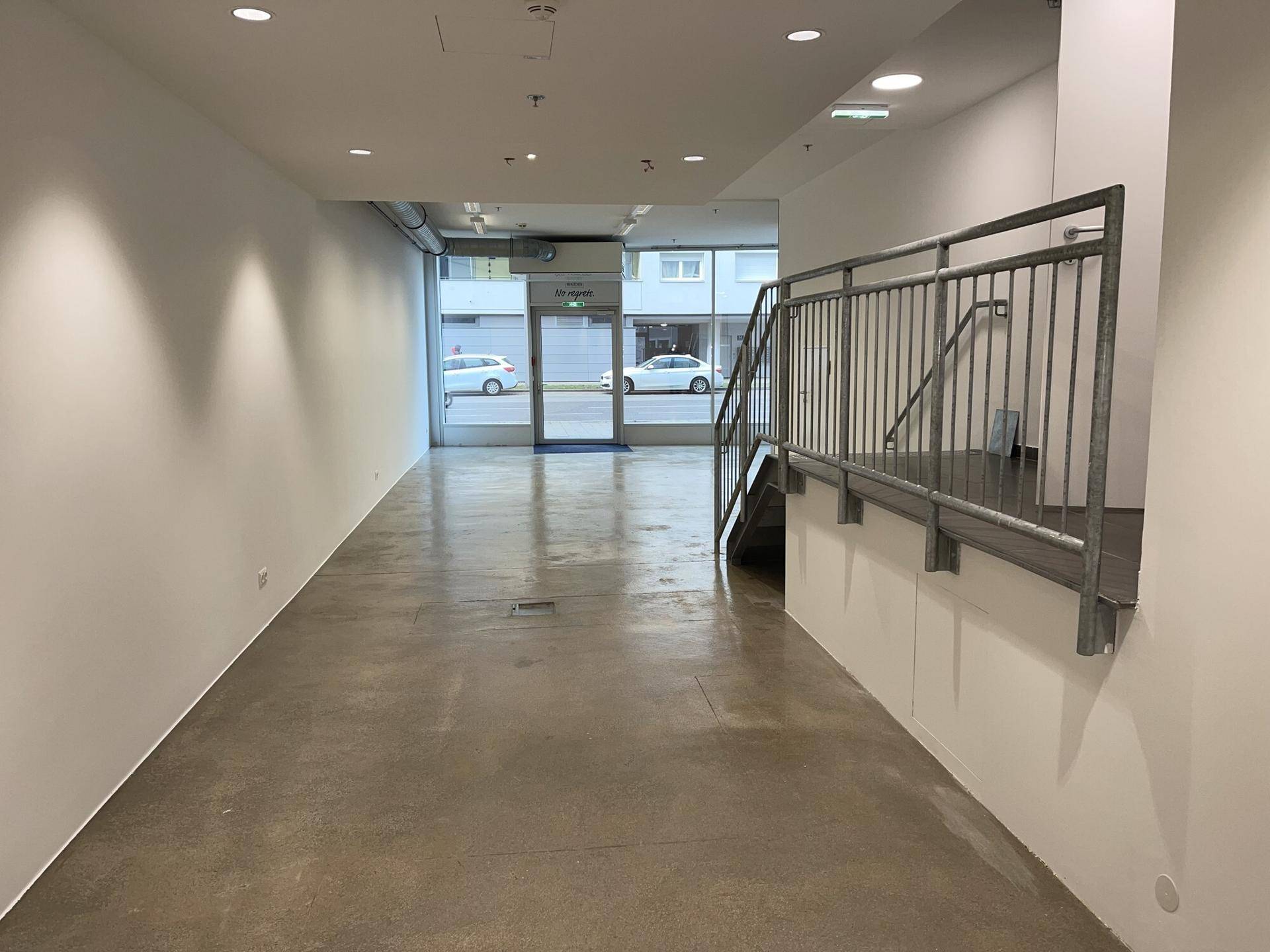 Leerer Gewerberaum mit poliertem Betonboden, großen Schaufenstern und einer Metalltreppe zum oberen Bereich.