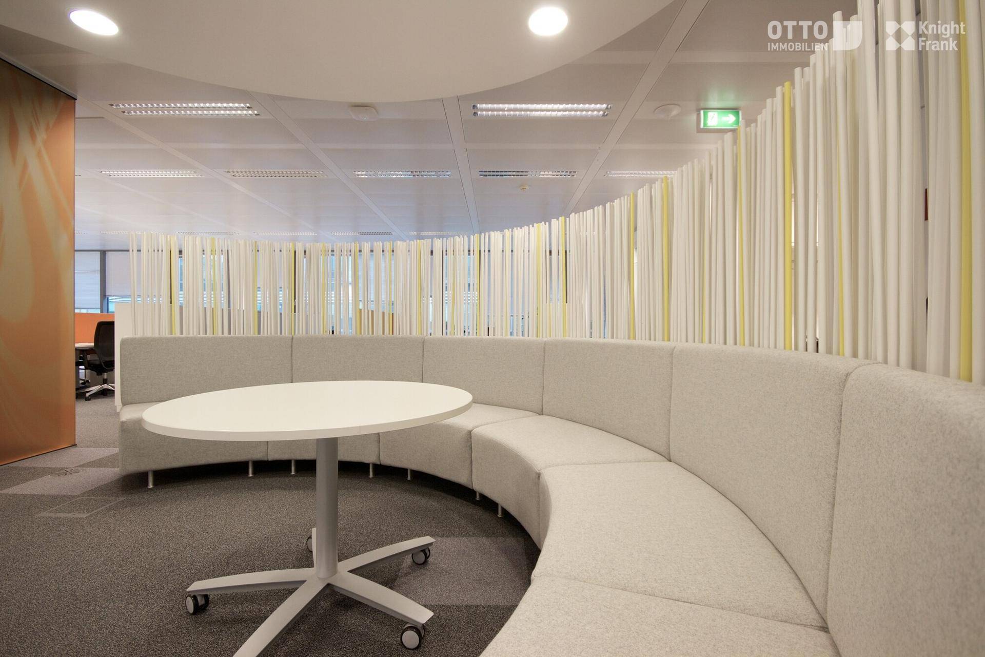 Moderner Bürobereich mit einer geschwungenen Sitzbank, einem runden Tisch und dekorativen Trennwänden.