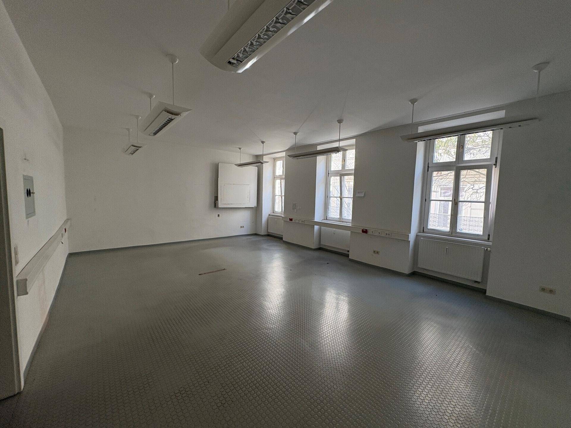 Heller Büroraum mit grauem Vinylboden und mehreren Fenstern, die den Raum gut belichten.