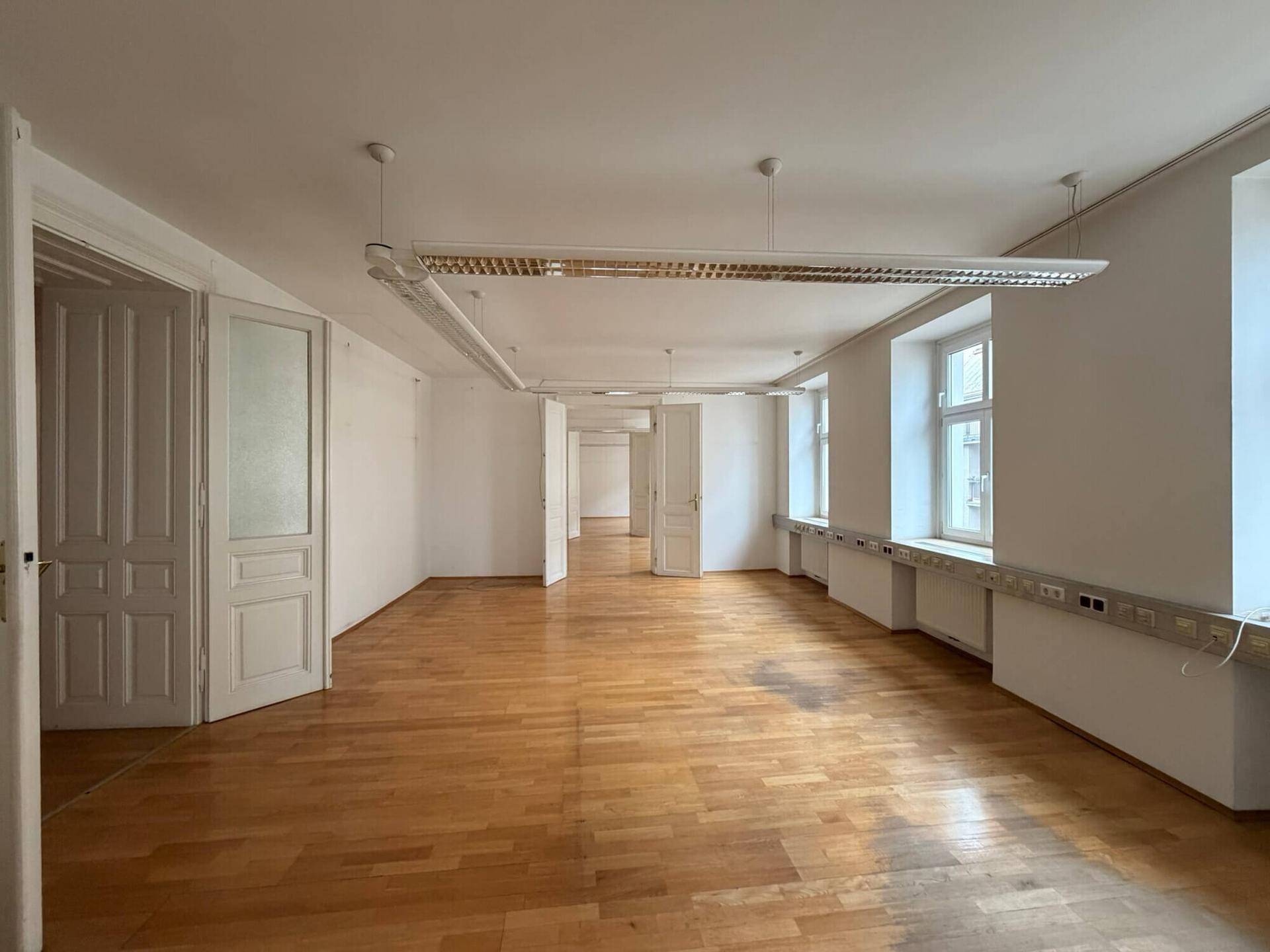 Langer Büroraum mit Holzboden, mehreren Fenstern und einer Reihe von Leuchtstoffröhren an der Decke für gute Beleuchtung.