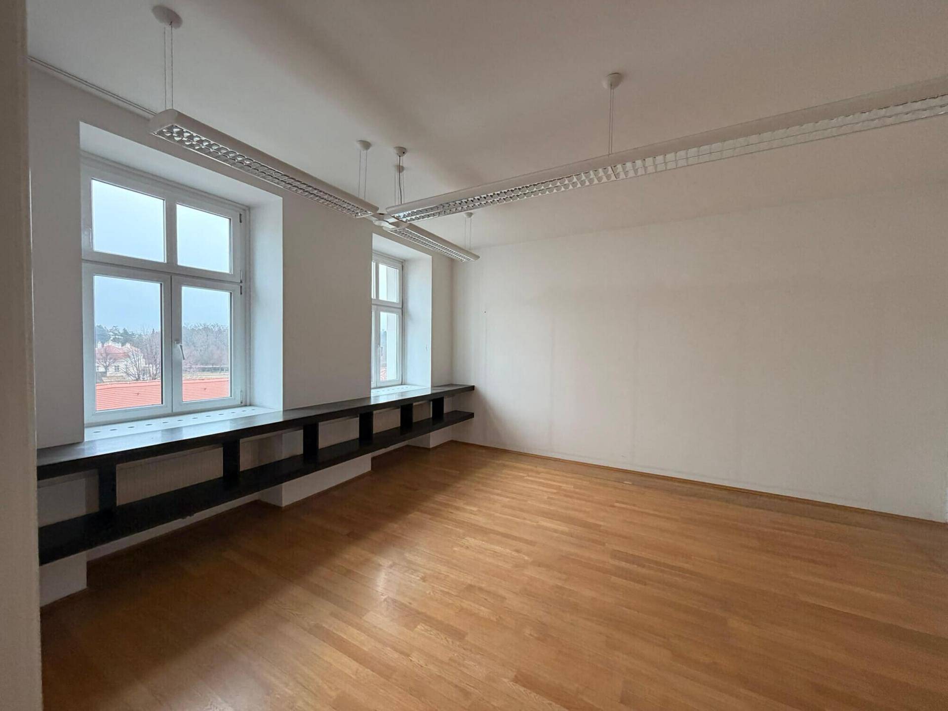 Heller Büroraum mit Holzboden, großen Fenstern und einer langen, dunklen Ablagefläche unter den Fenstern.