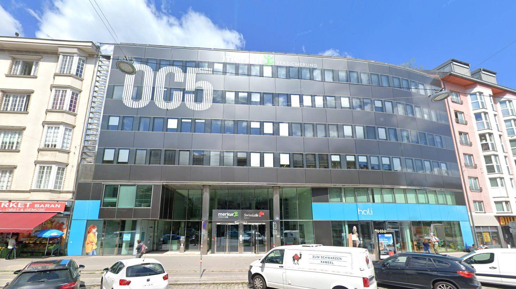 Modernes Bürogebäude mit dunkler Fassade, großen Fenstern und Geschäften im Erdgeschoss an einer belebten Straße.