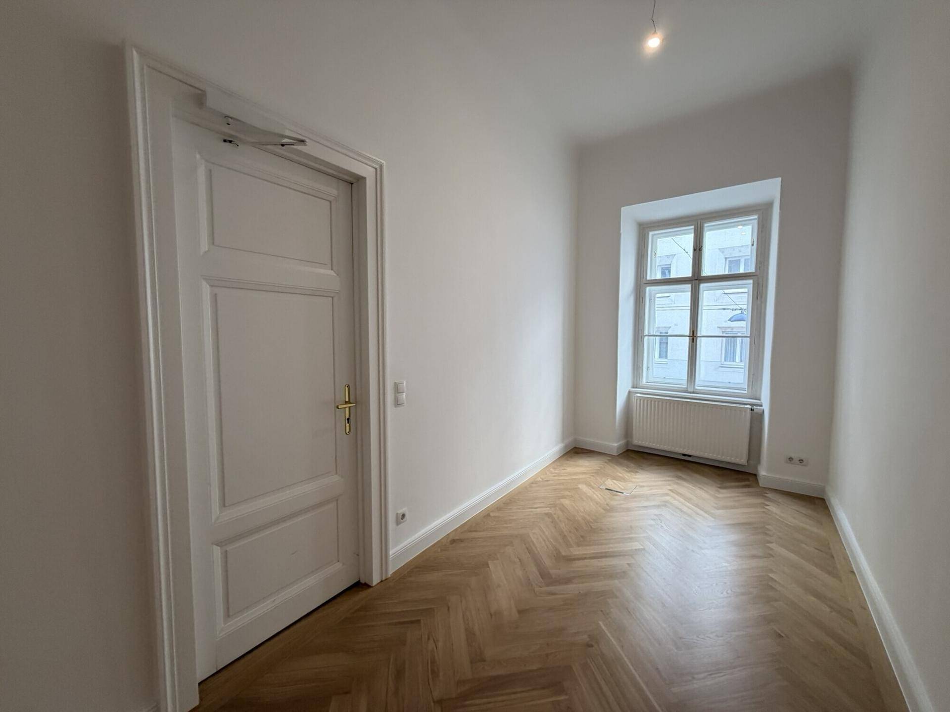 Helles Zimmer mit Fischgrätparkett und einer klassischen Tür, ideal als Schlaf- oder Arbeitszimmer.