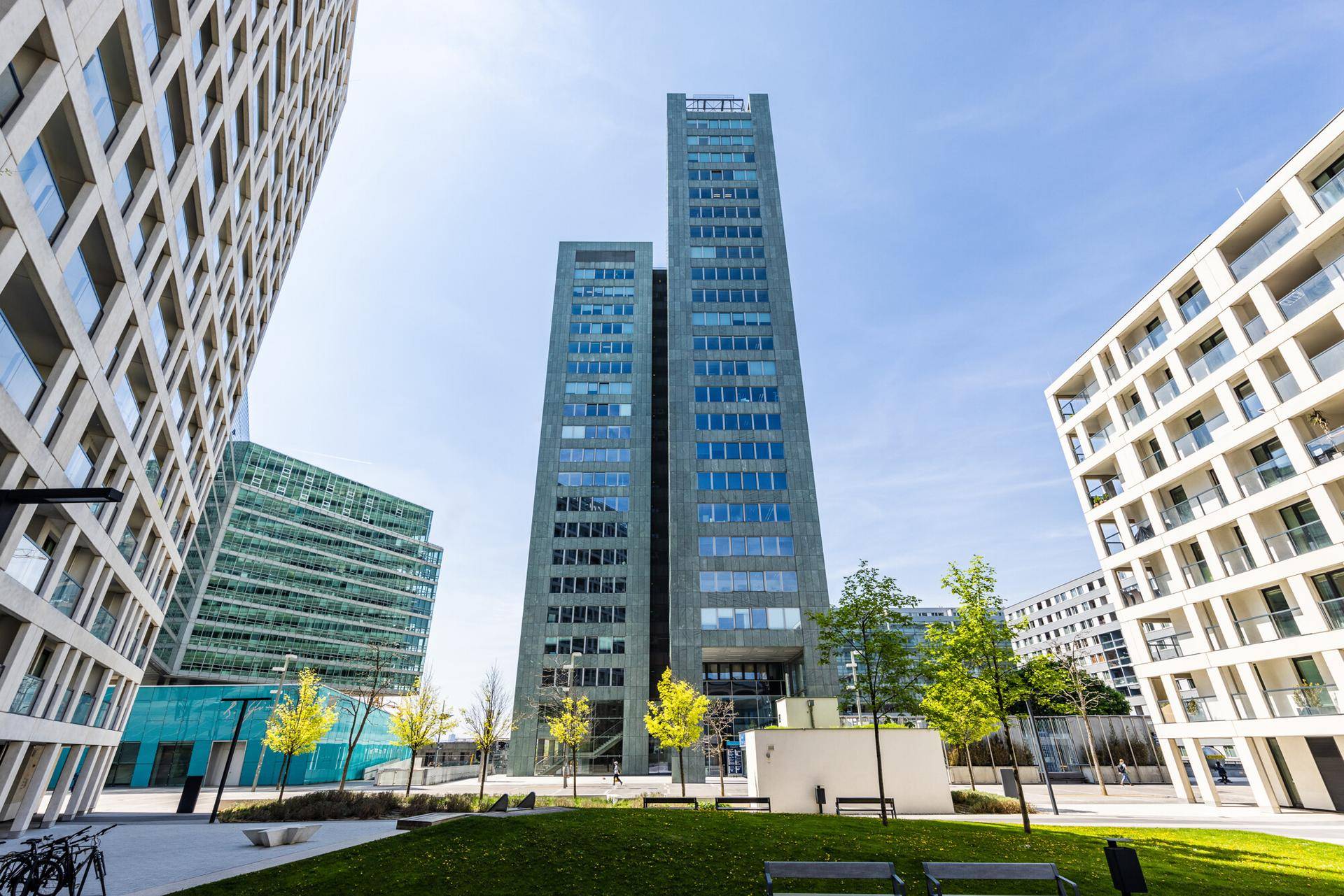 Moderner Wolkenkratzer umgeben von anderen Gebäuden und einer gepflegten Grünfläche mit Bänken.