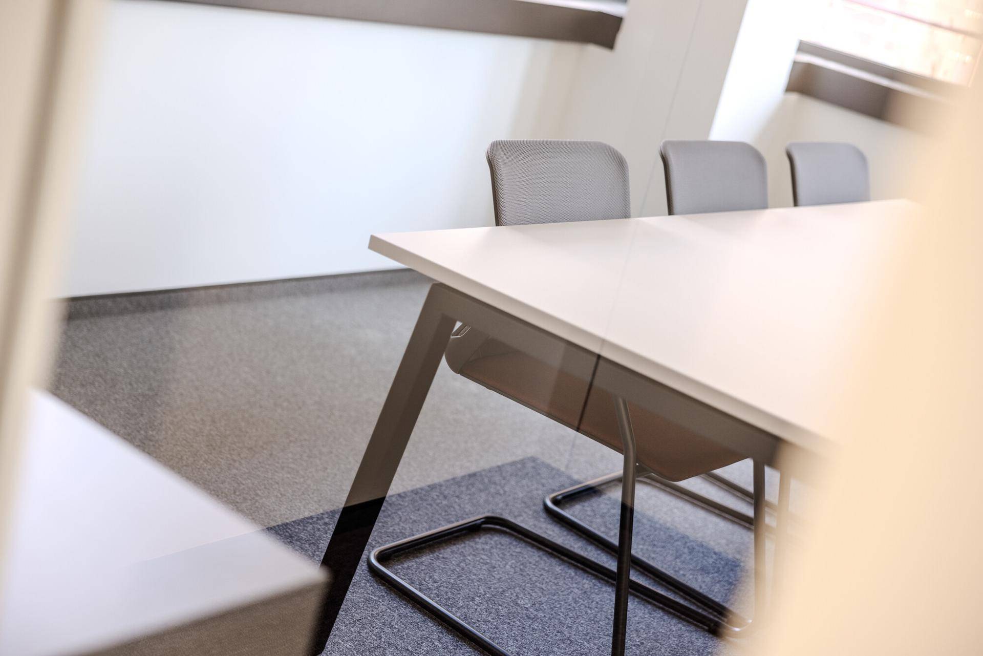 Nahaufnahme eines modernen Bürotisches mit grauen Stühlen und Teppichboden in einem hellen Raum.