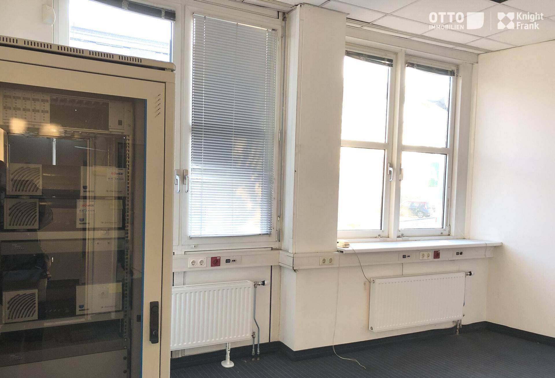 Heller Büroraum mit großen Fenstern, Heizkörpern und einem Serverschrank, ideal für Verwaltungsbereiche.