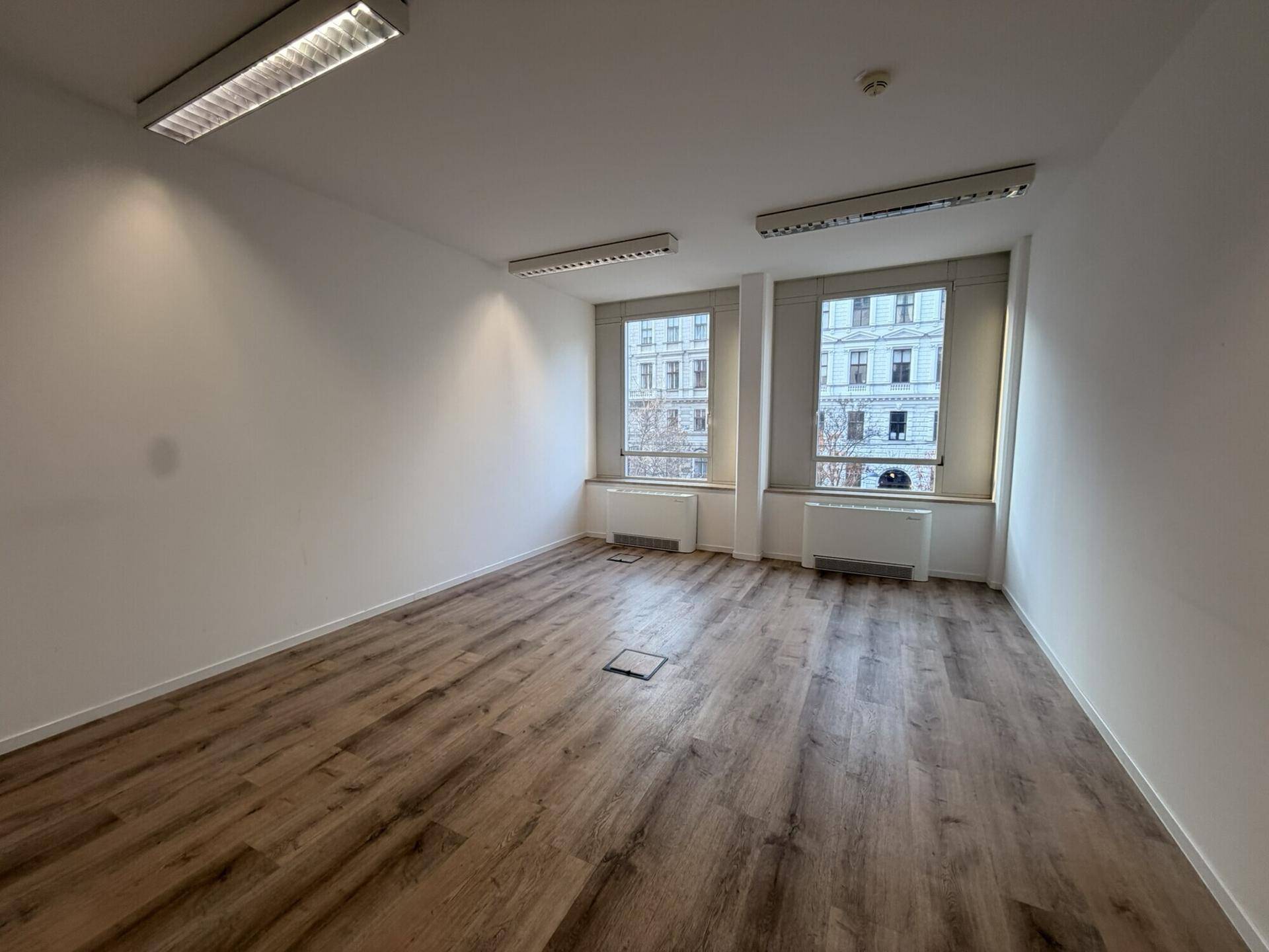 Leerer Büroraum mit Laminatboden und zwei Fenstern, die einen Blick auf die Stadt bieten.