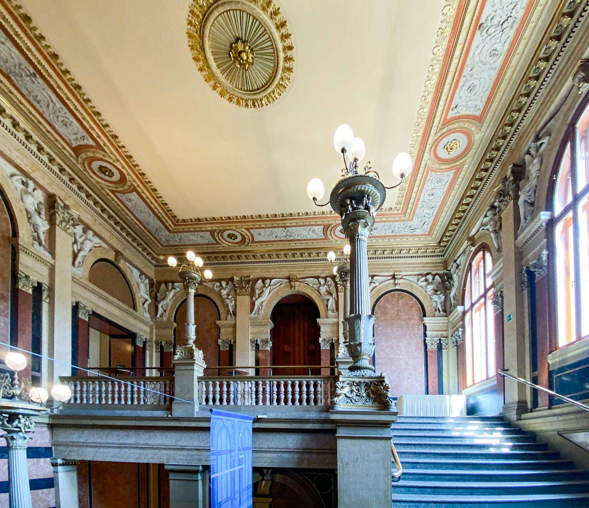 Beeindruckendes Treppenhaus mit kunstvollen Verzierungen, hohen Decken und großen Fenstern.
