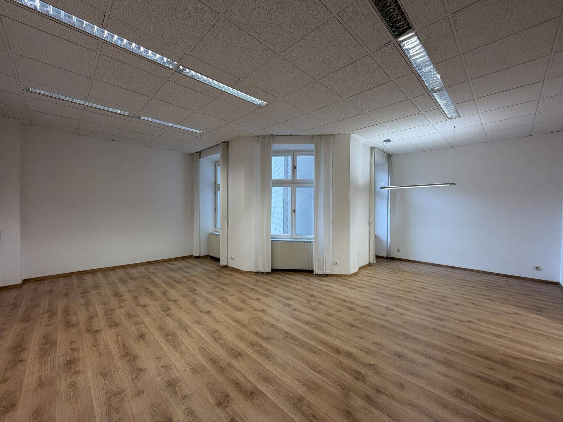 Geräumiges Büro mit Laminatboden, großen Fenstern und Vorhängen, bereit zur individuellen Einrichtung.
