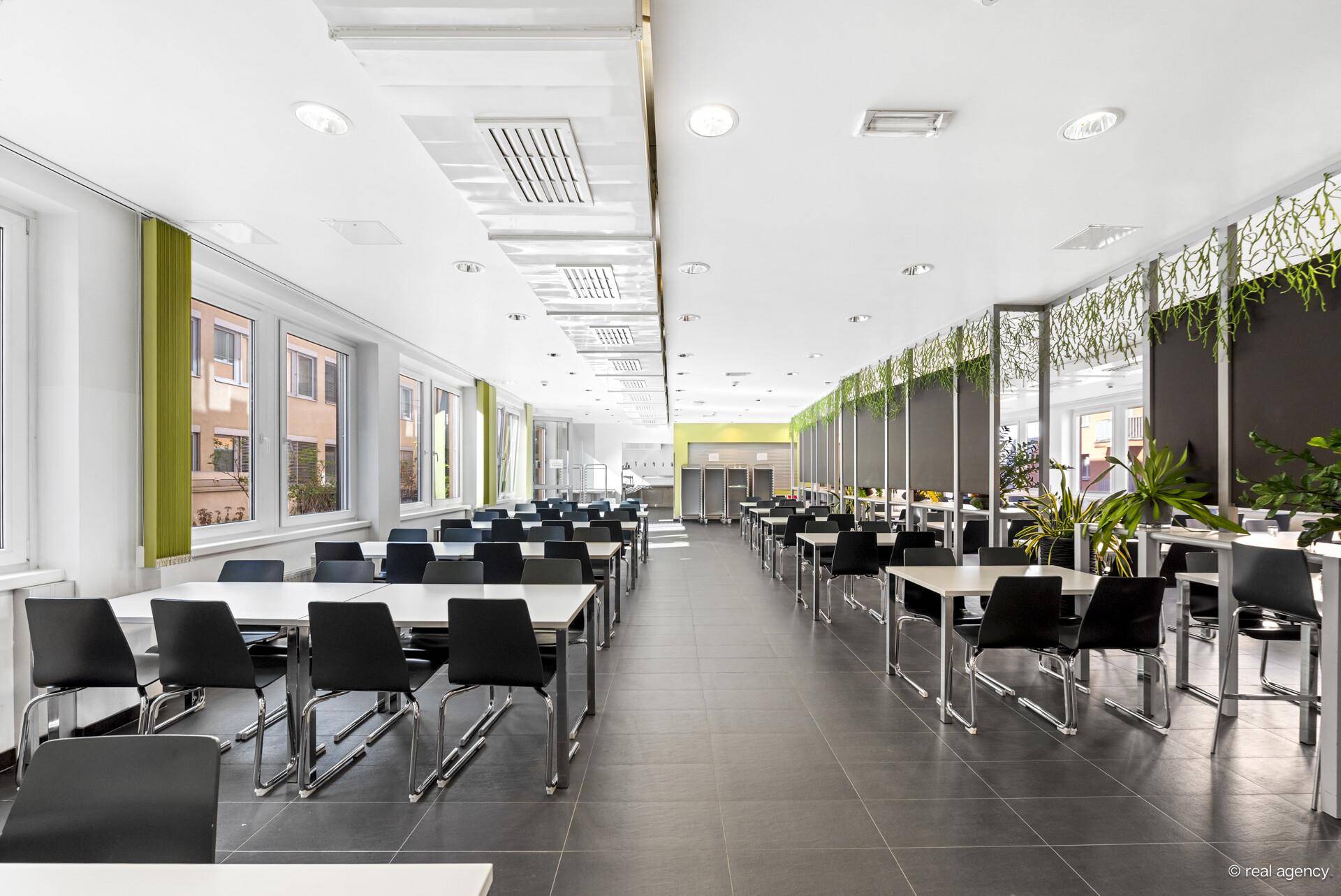 Große, helle Kantine mit vielen Tischen und Stühlen, großen Fenstern und grünen Dekorationen.