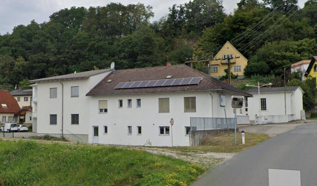 Gebäude mit weißer Fassade und Solarmodulen auf dem Dach, eingebettet in eine grüne Hügellandschaft.