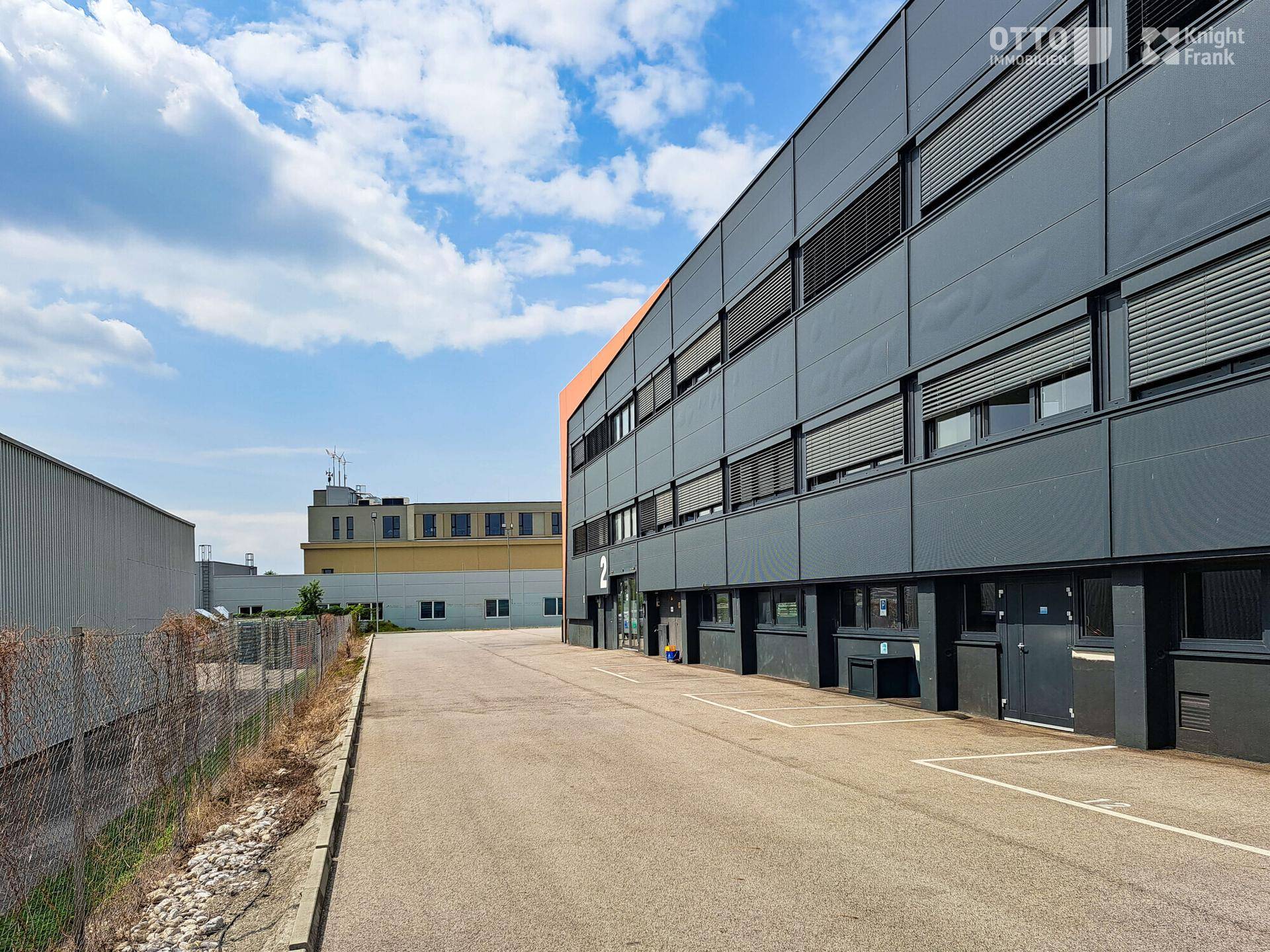 Modernes Gewerbegebäude mit dunkler Fassade, vielen Fenstern und einem großen Parkplatz davor unter blauem Himmel.
