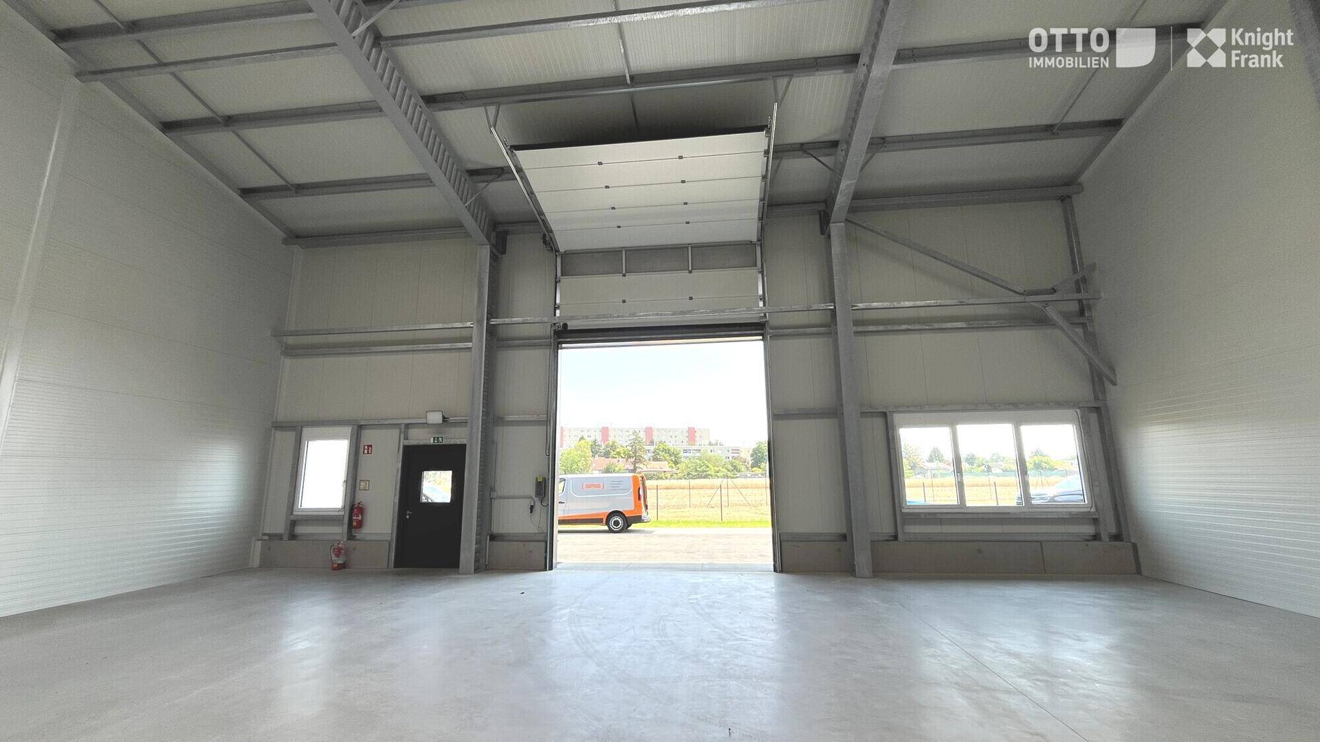 Geräumige Lagerhalle mit hohen Decken, großem Rolltor und viel Tageslicht durch Fenster.