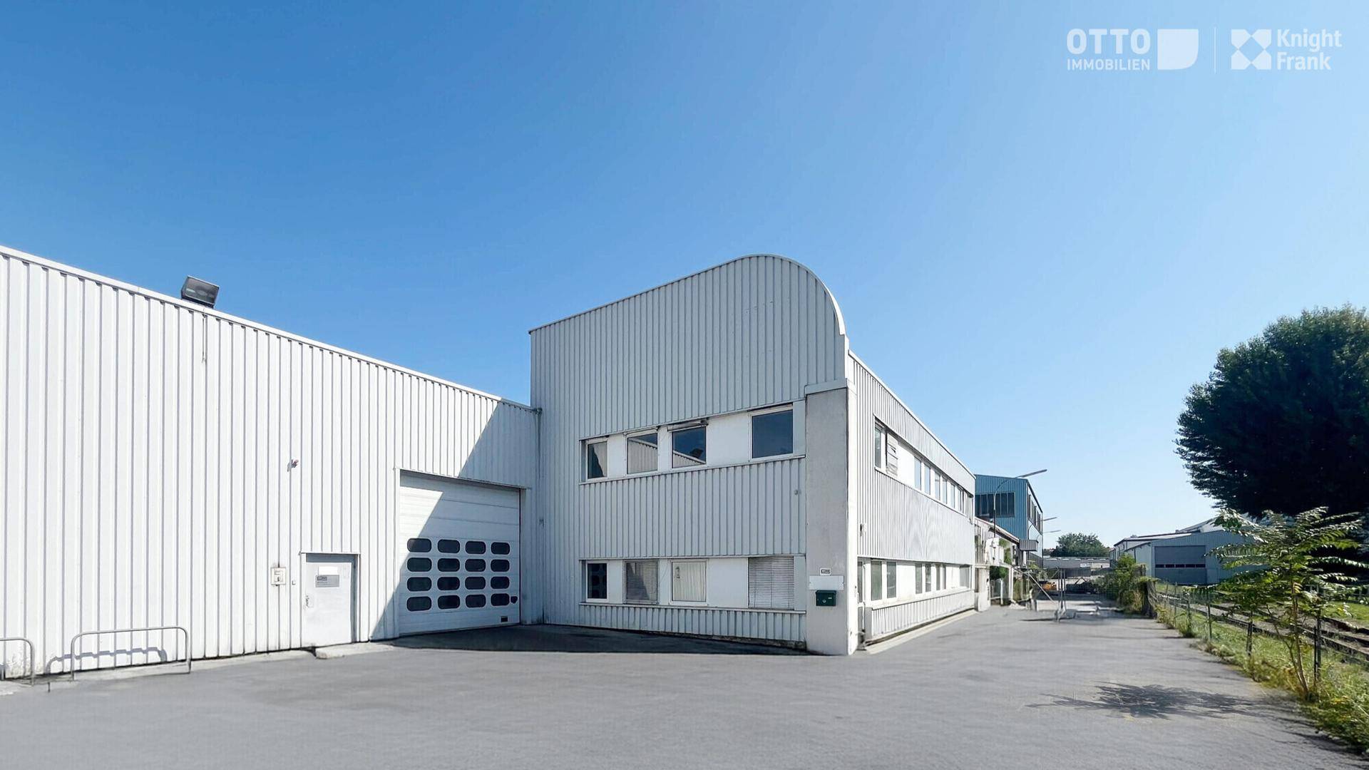 Außenansicht eines Industriegebäudes mit heller Wellblechfassade und großem Rolltor unter blauem Himmel.