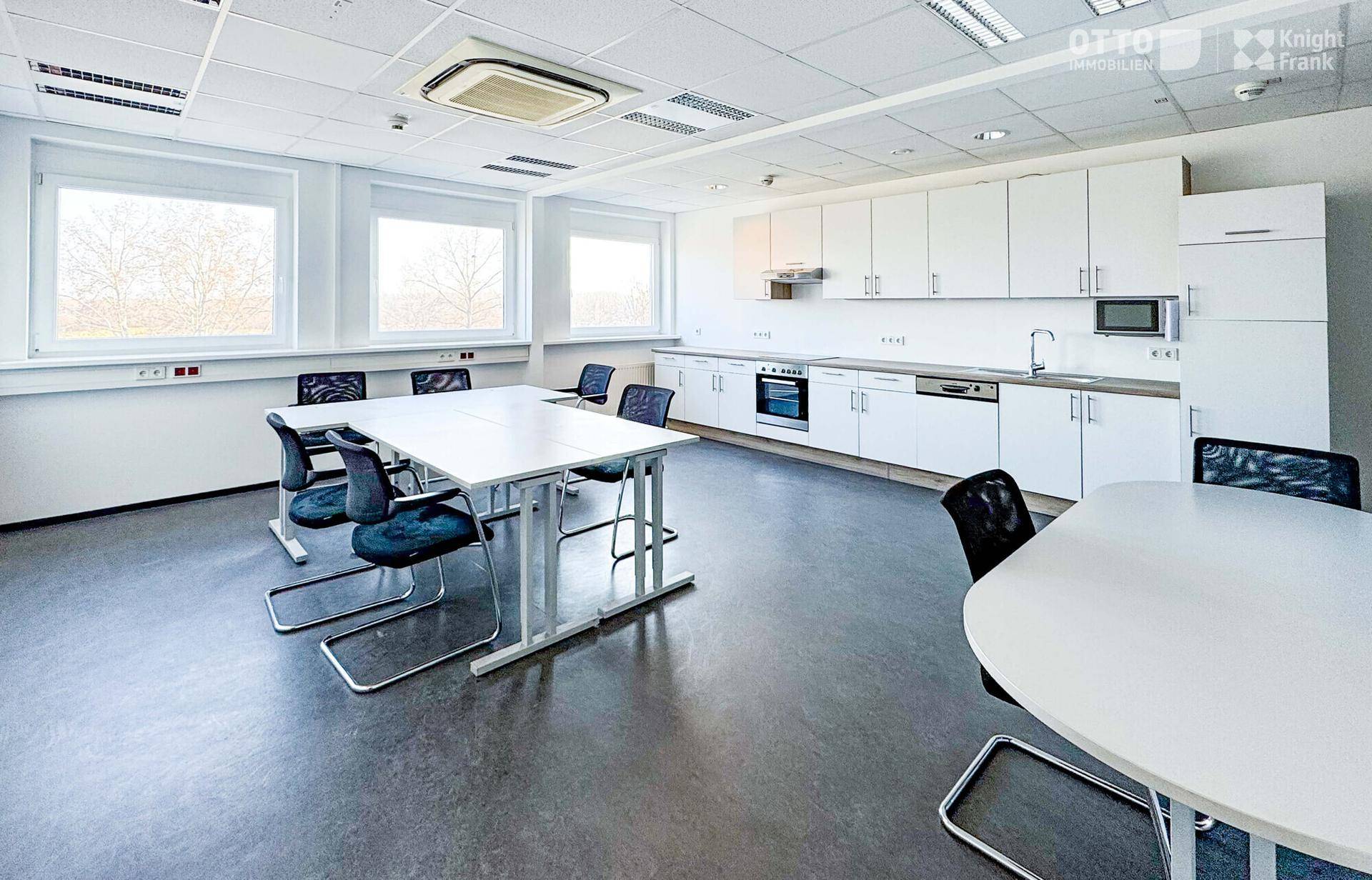 Moderne Büroküche mit weißen Schränken, Esstischen und großen Fenstern für viel Tageslicht.