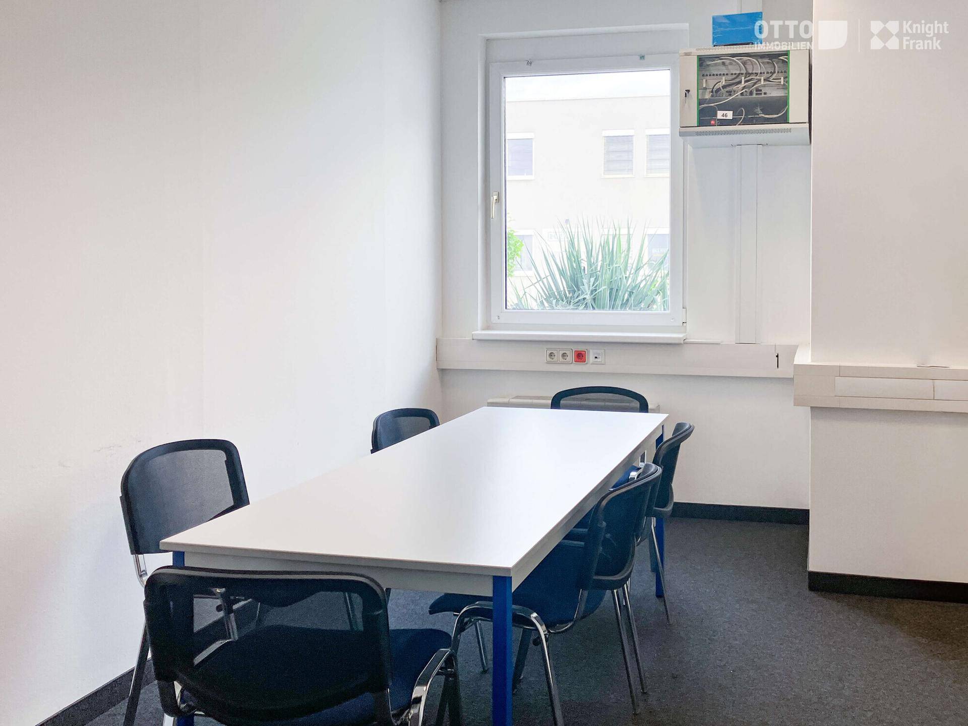 Kleiner Besprechungsraum mit einem weißen Tisch, blauen Stühlen und einem Fenster mit Blick ins Grüne.