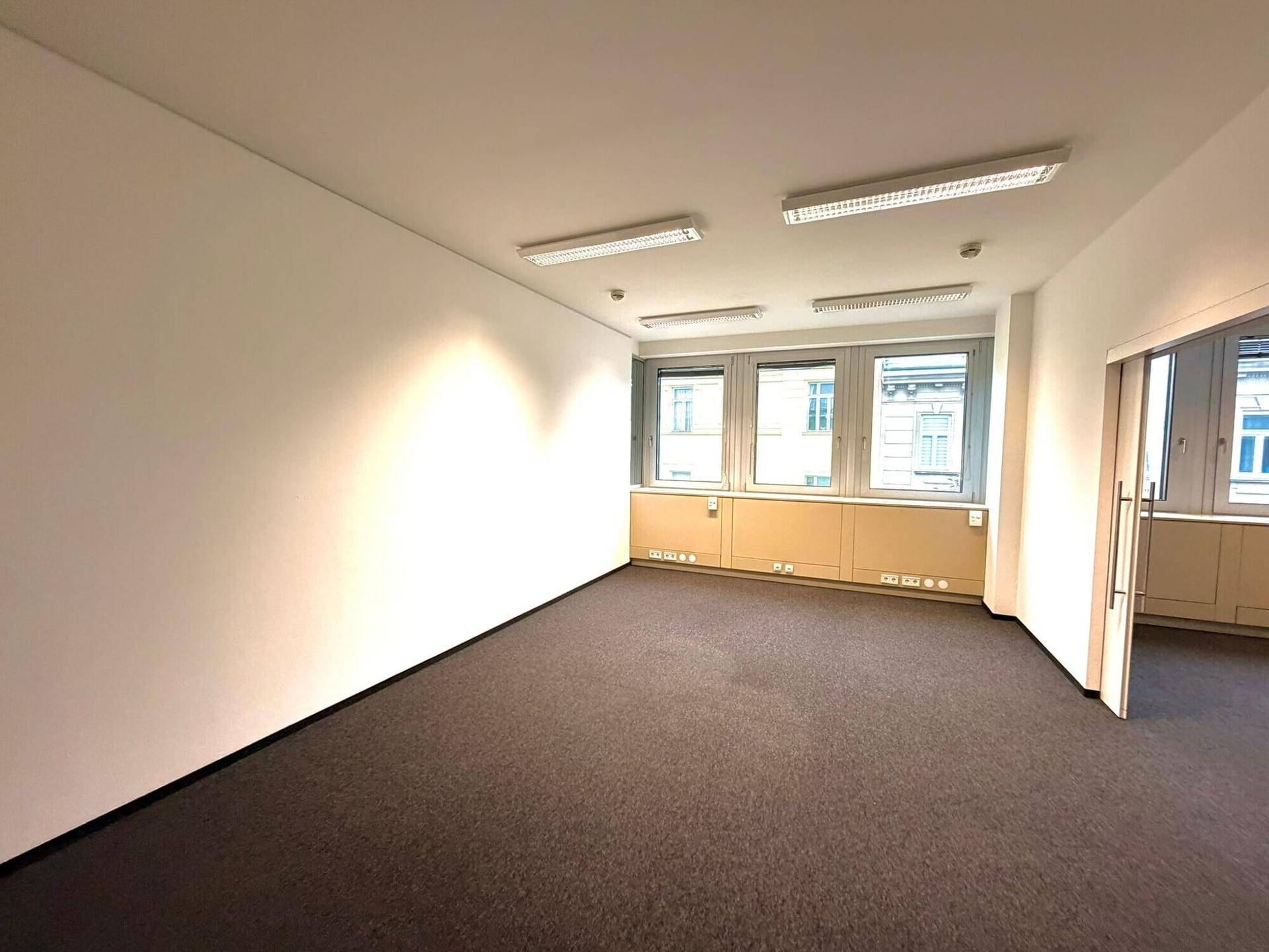 Heller Büroraum mit Teppichboden, großen Fenstern und ausreichend Steckdosen entlang der Wand.