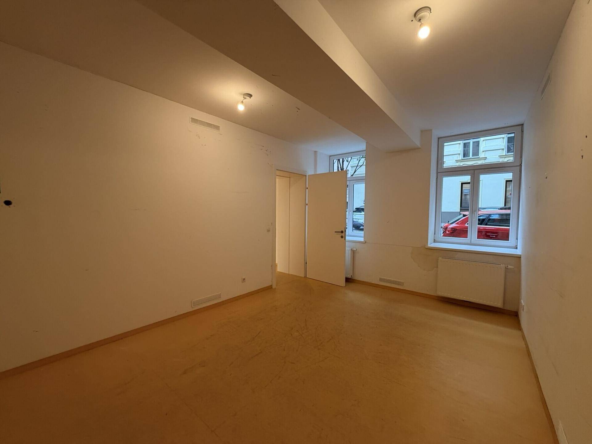 Heller, leerer Raum mit Fenstern zur Straße, weißen Wänden und gelbem Boden, geeignet als Büro.