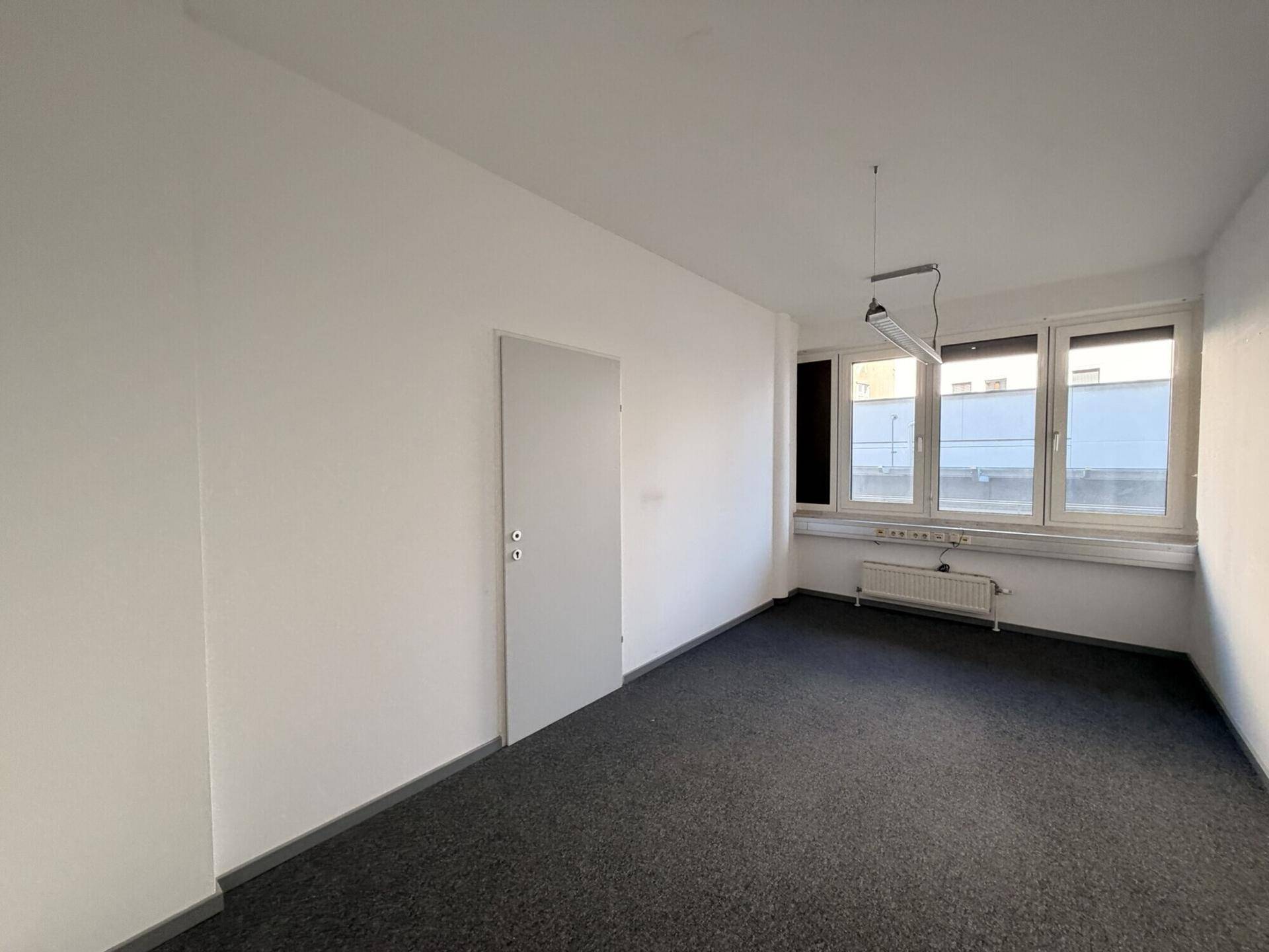Schlichtes, leeres Büro mit dunklem Teppichboden, weißer Tür und Fenstern, die Tageslicht spenden.