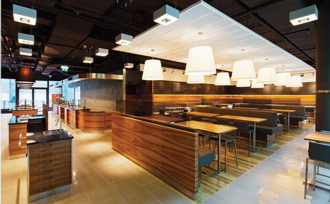 Moderne Cafeteria mit Holzelementen, verschiedenen Sitzbereichen und einer langen Theke für Speisen und Getränke.